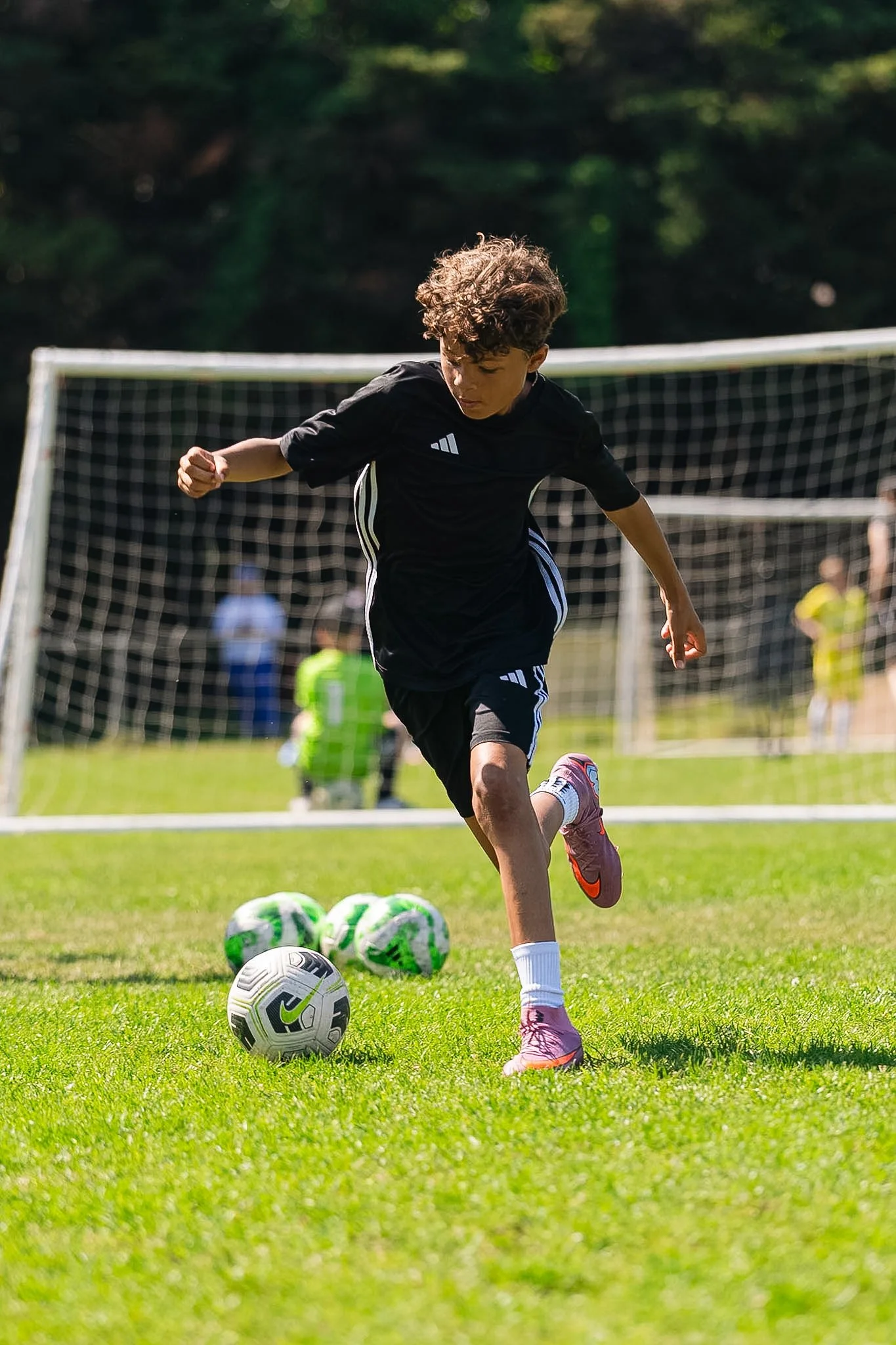 Young boy playing soccer on a grassy field during daytime, with soccer balls in front of him and goalposts in the background.