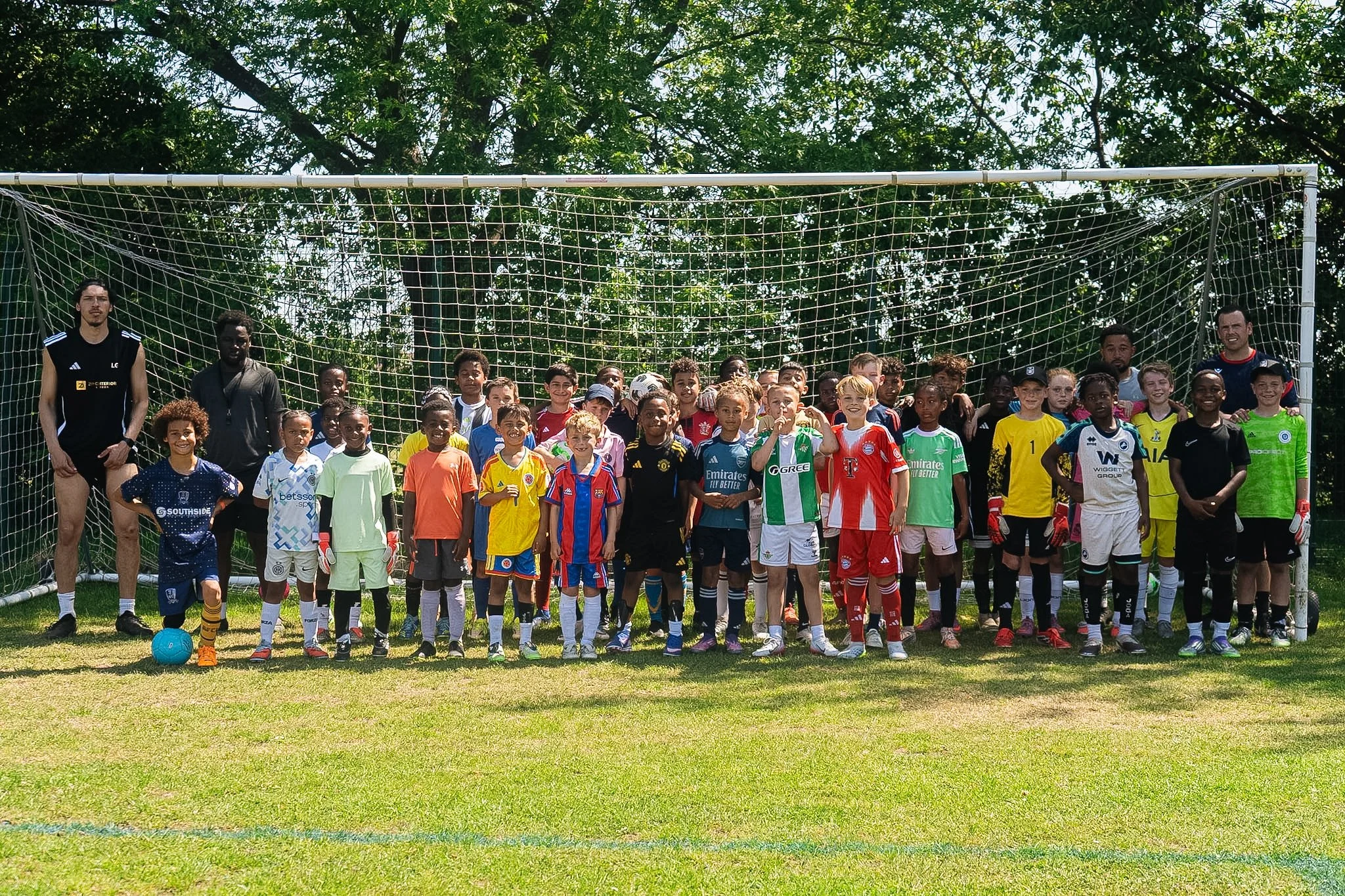 Group of children and adults standing in front of a soccer goal on a grassy field, with trees in the background, during daytime.