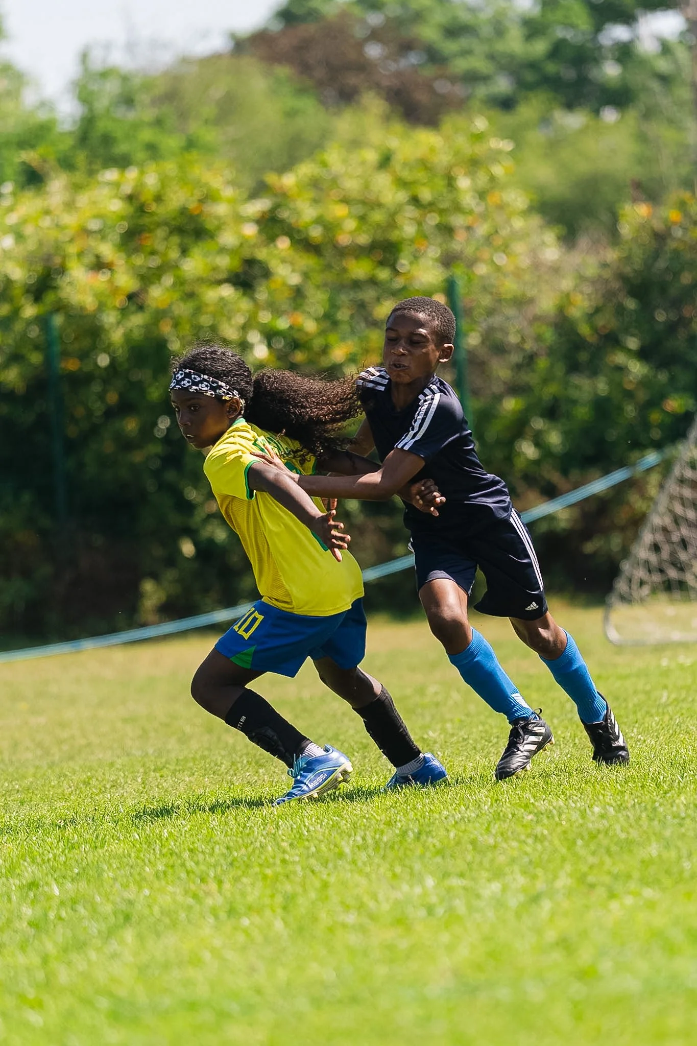 Two young boys are engaged in a soccer match outdoors on a grassy field, with trees and greenery in the background, both running and competing for the ball.