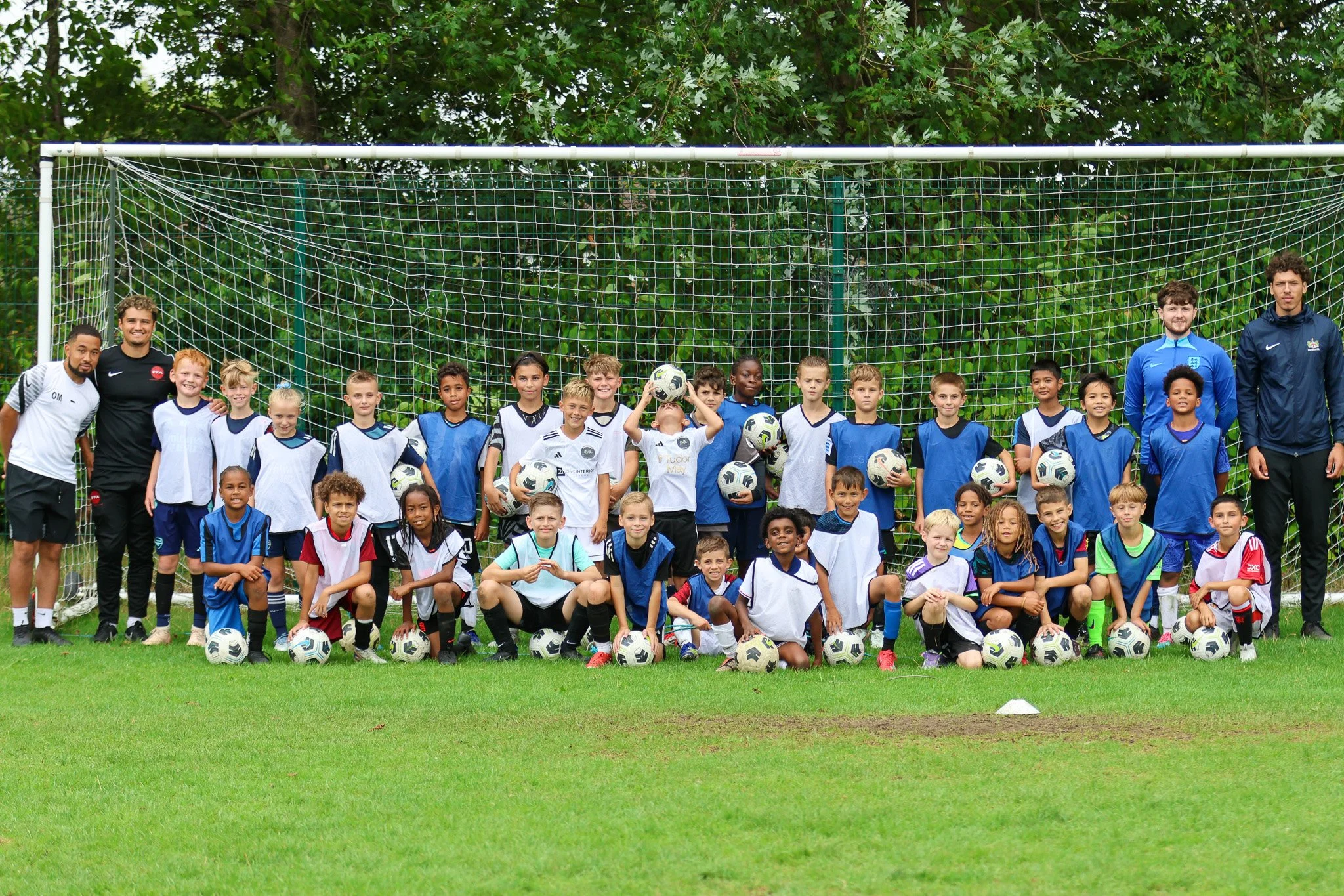 Group photo of young soccer players and coaches in front of a goalpost on a grassy field, some children holding soccer balls, with lush green trees in the background.