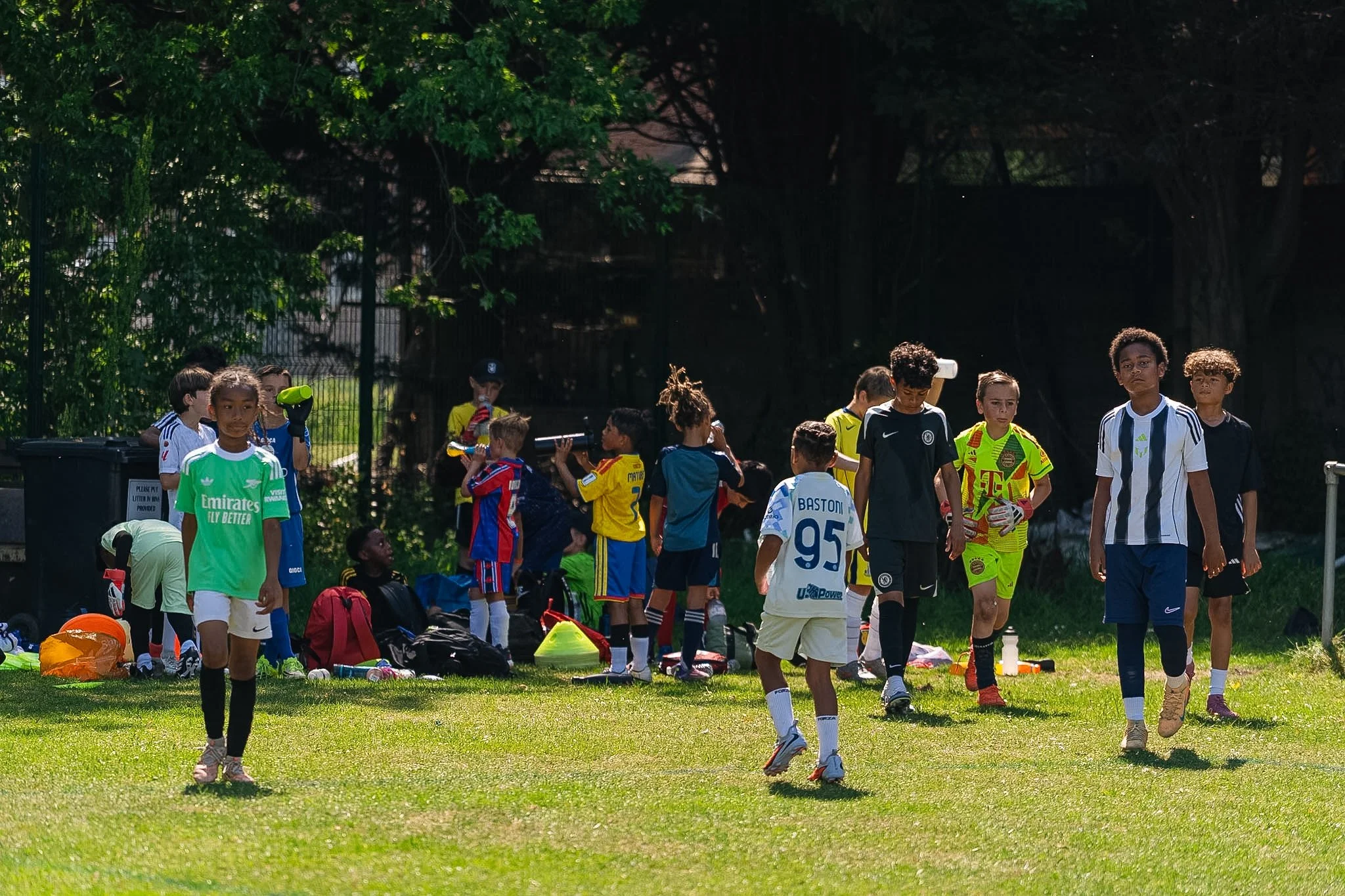 Children and teens in soccer uniforms gather on a grassy field after practice or a game, with some holding sports equipment, others standing or walking, and trees in the background.