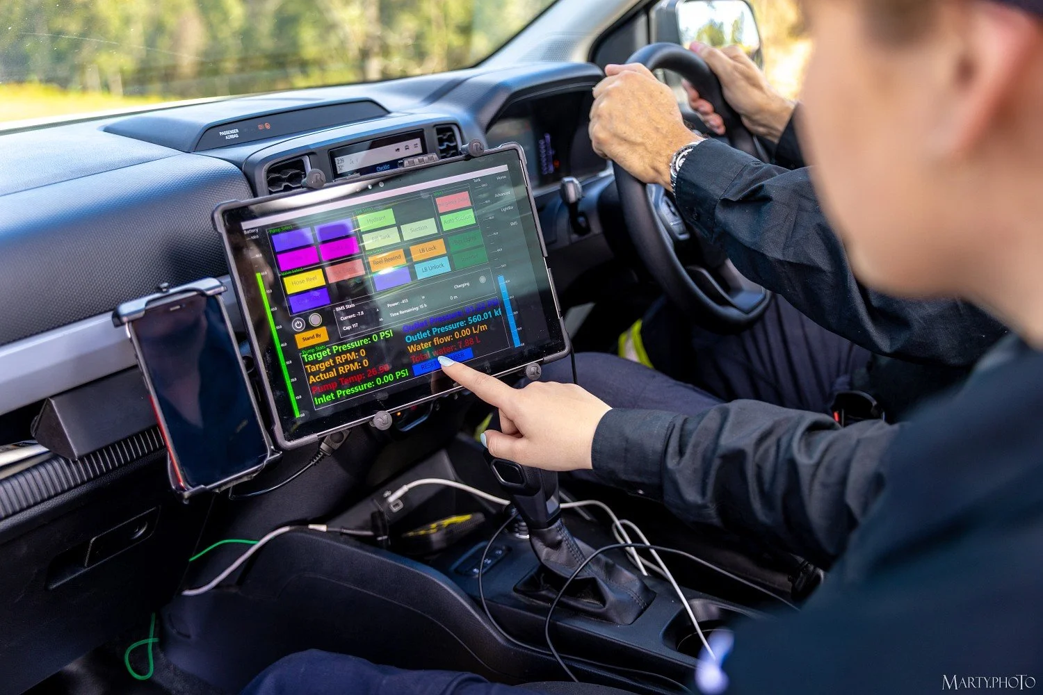Person in a car operating a digital control panel for a vehicle's systems, with another person in the driver's seat.