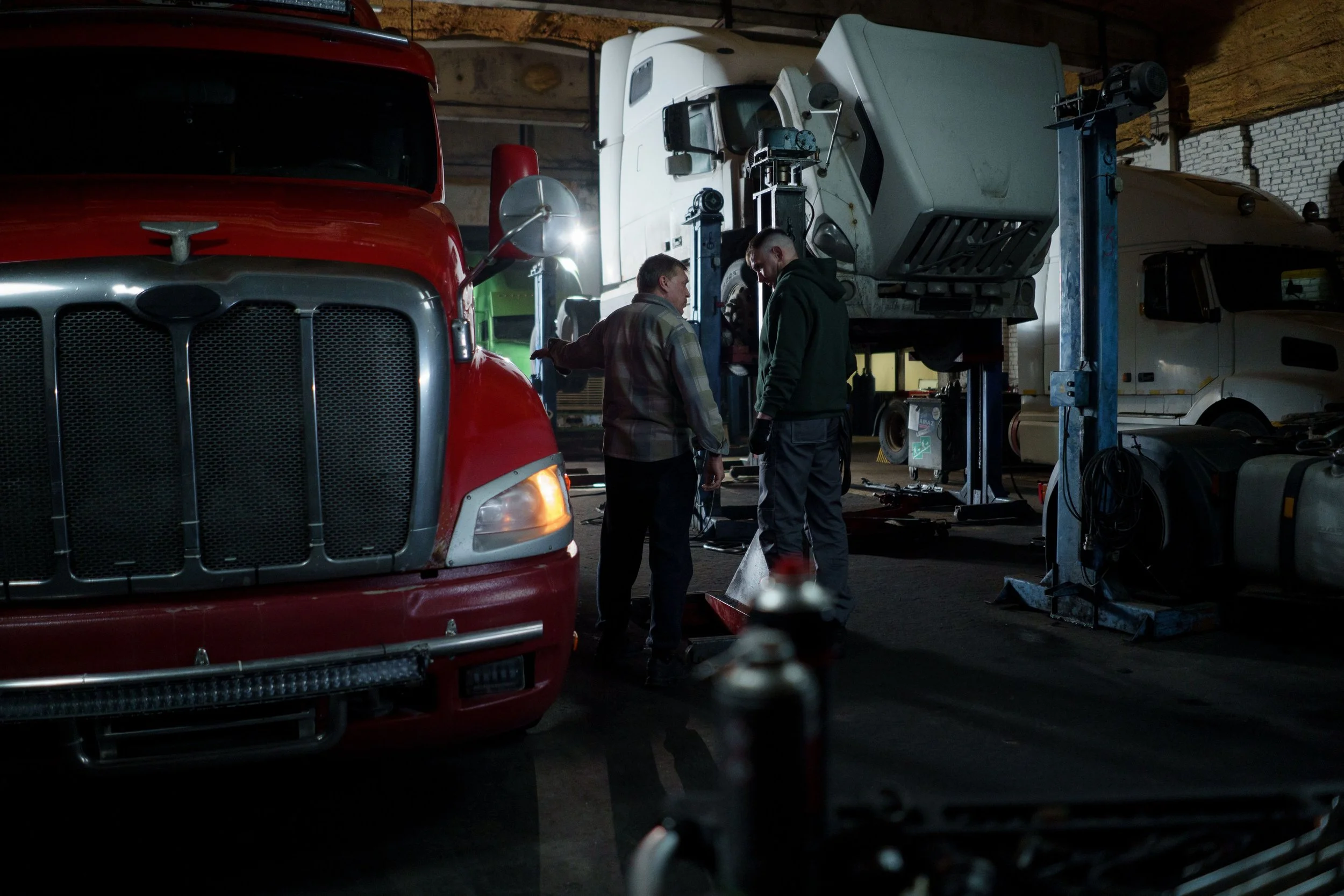 Two men working on a large white truck inside a dimly lit garage, with a red semi-truck visible in the foreground and other vehicles and equipment around.