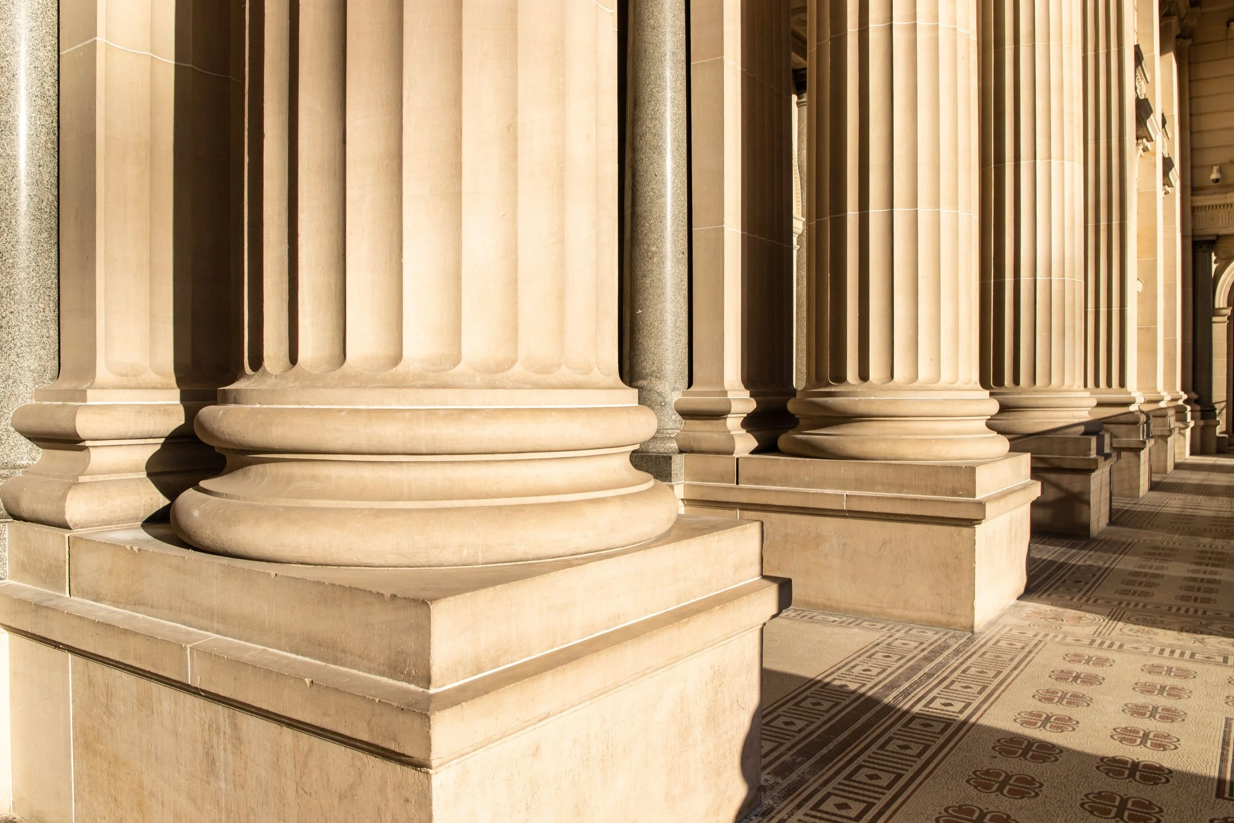 Close-up of large stone columns with fluted shafts and rounded bases, part of a classical building façade. Shadows cast by the columns fall on the tiled floor.