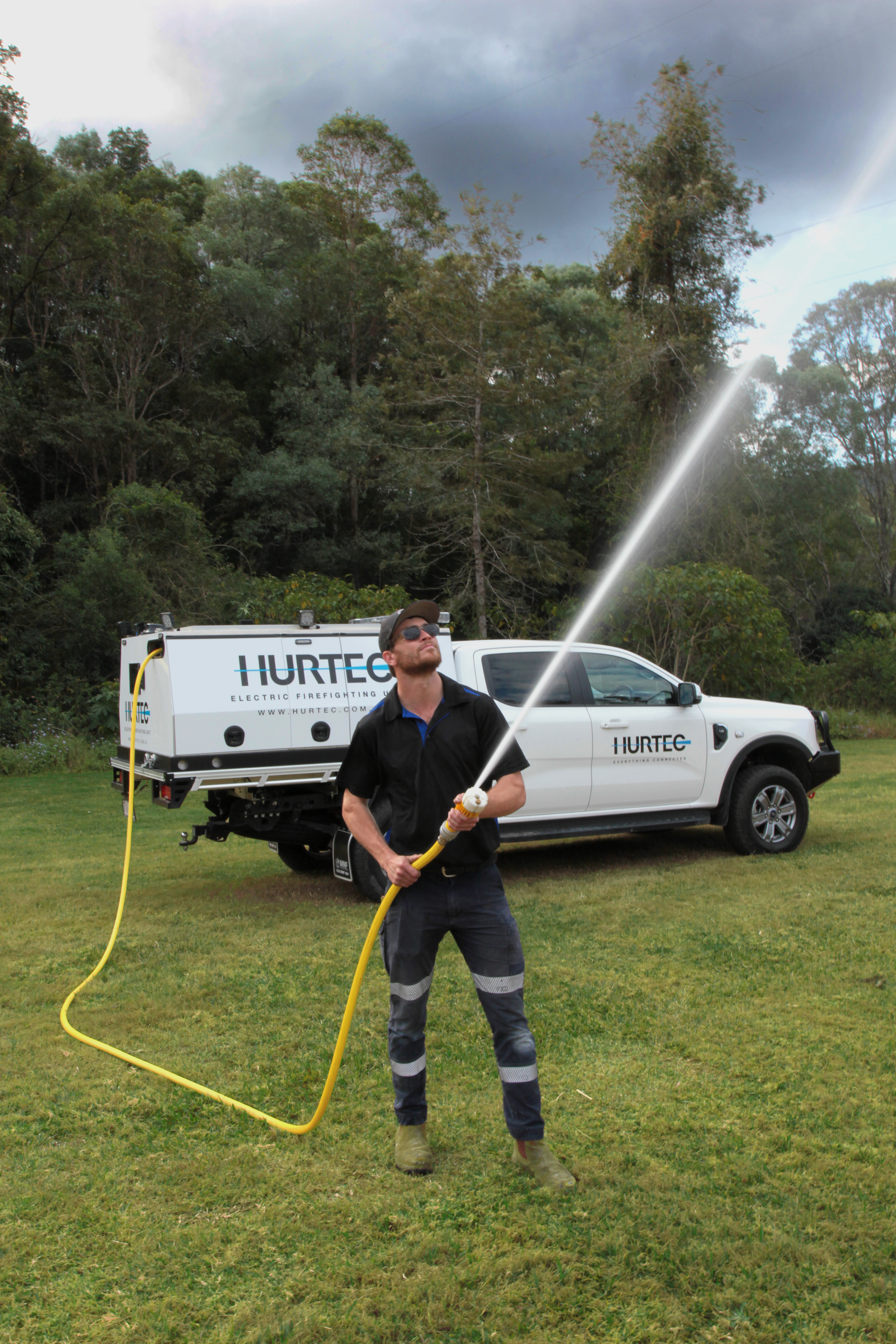 A man with a beard and sunglasses holding a yellow hose in a grassy area with trees in the background, and a white truck labeled 'HURTEC' parked behind him.
