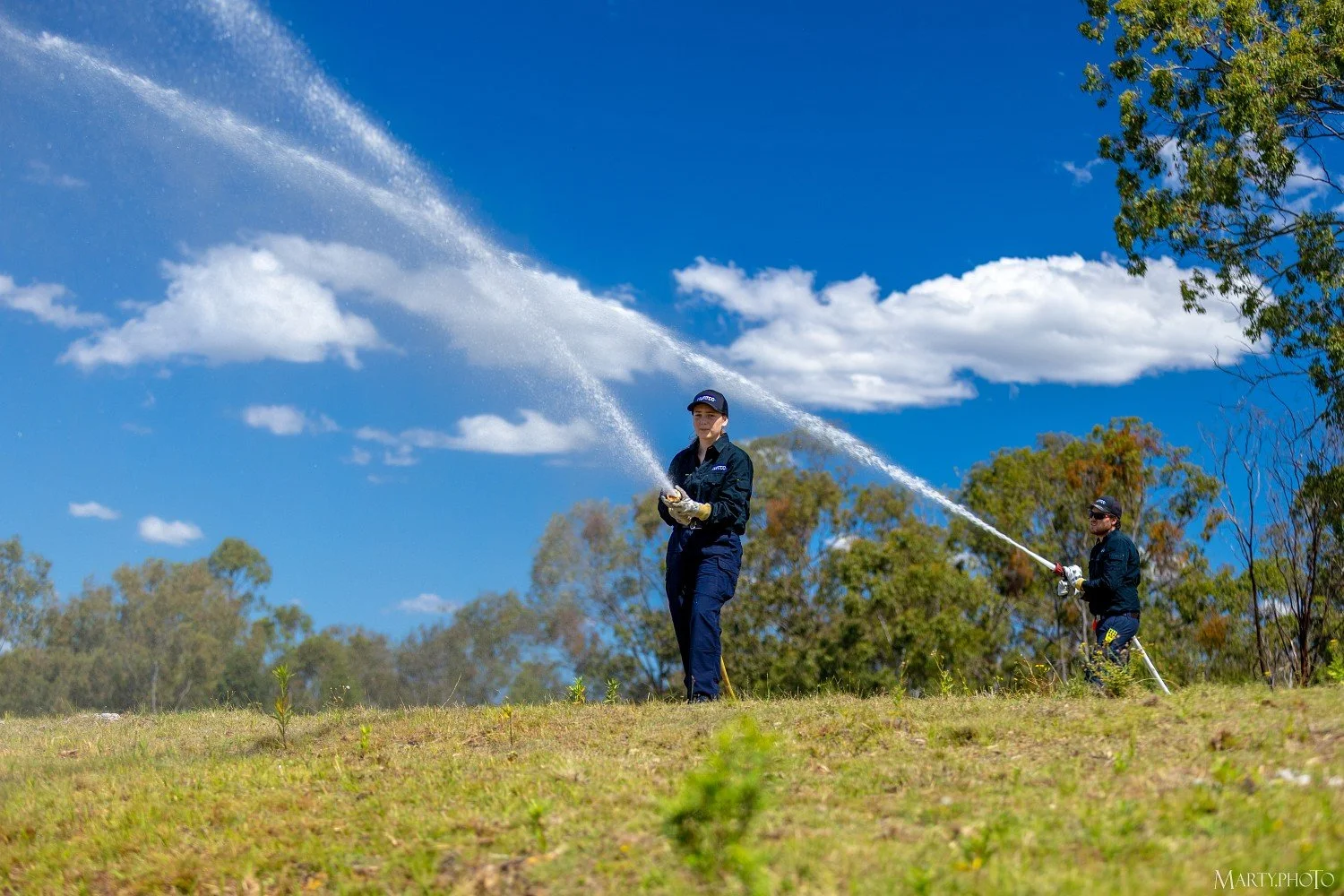 Two firefighters in navy uniforms and gloves watering grass outdoors under a bright blue sky with white clouds; one in the foreground and one in the background holding hoses fed by powerful Hurtec electric firefighting pumps.