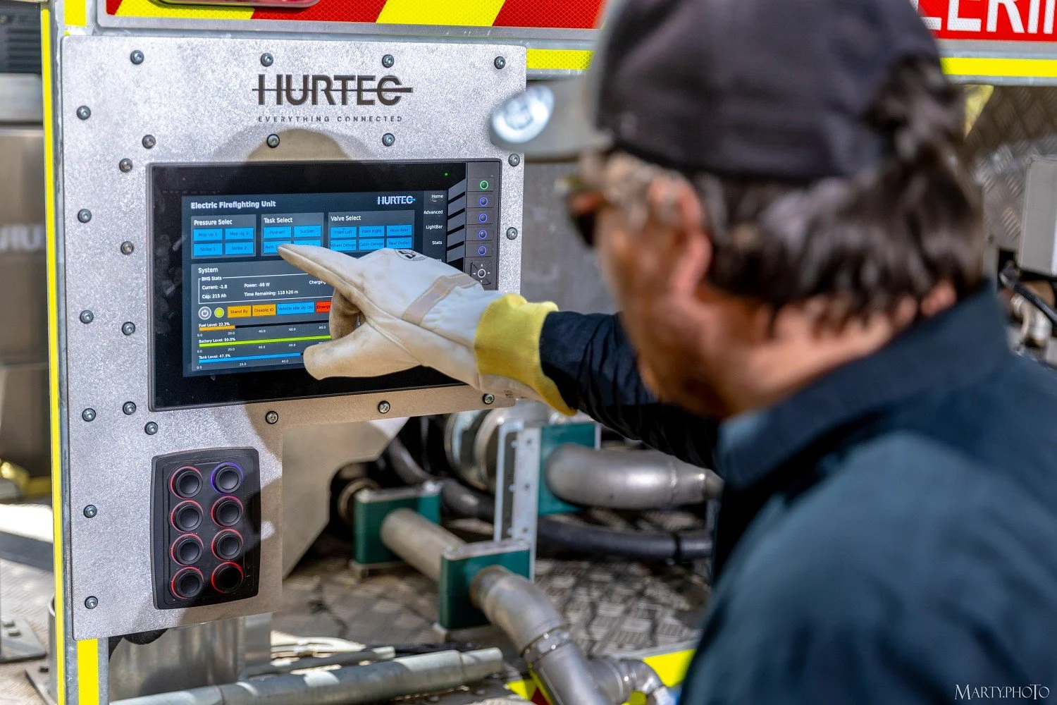 A worker wearing safety gloves and safety glasses operating an electric firefighting unit control panel mounted on a metal case labeled 'HURTEC.' The panel displays system status, pressure, and other operational metrics.