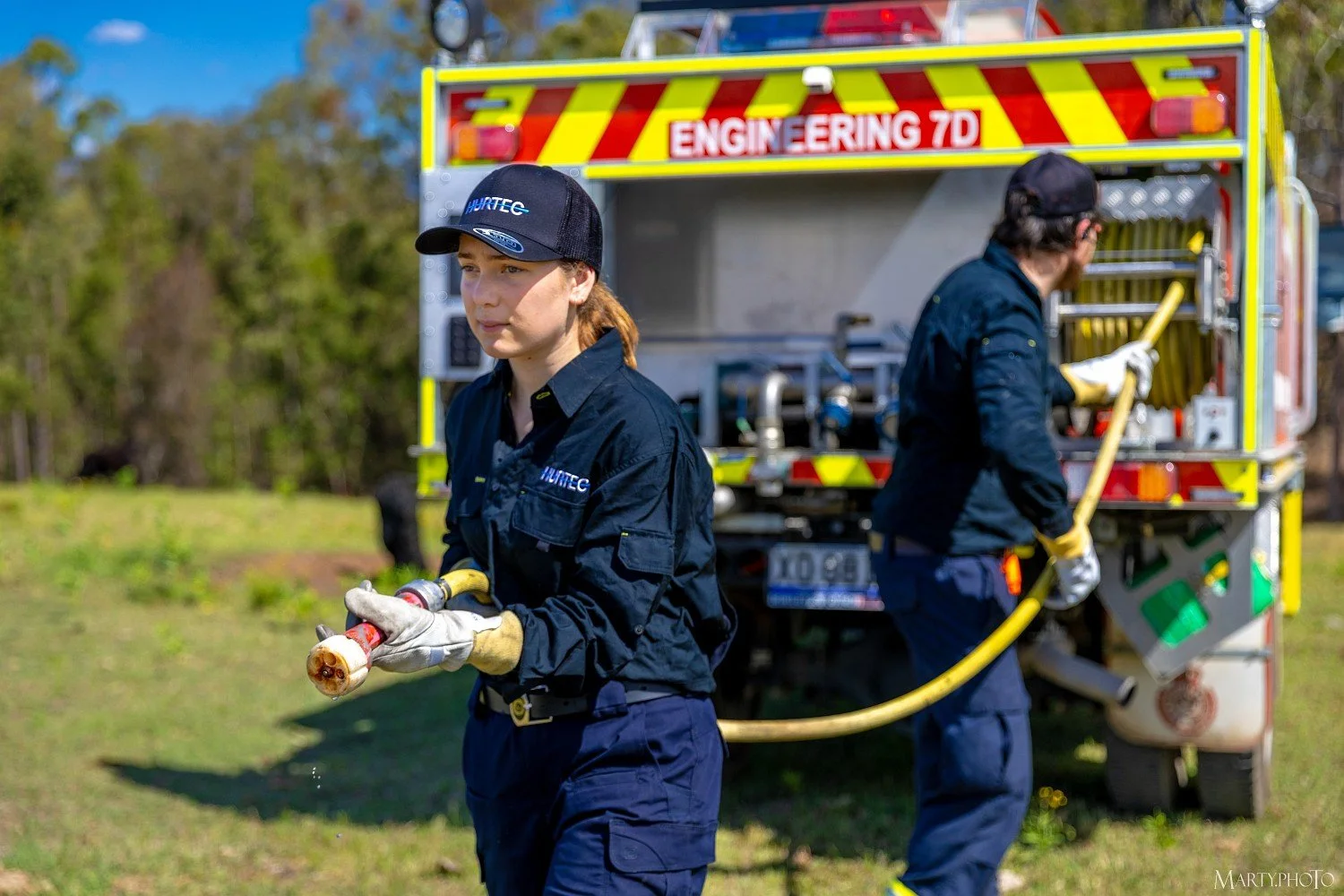 Two female firefighters in uniform handling a hose with a fire truck in the background. One is looking toward the camera, and the other is facing away, adjusting equipment.