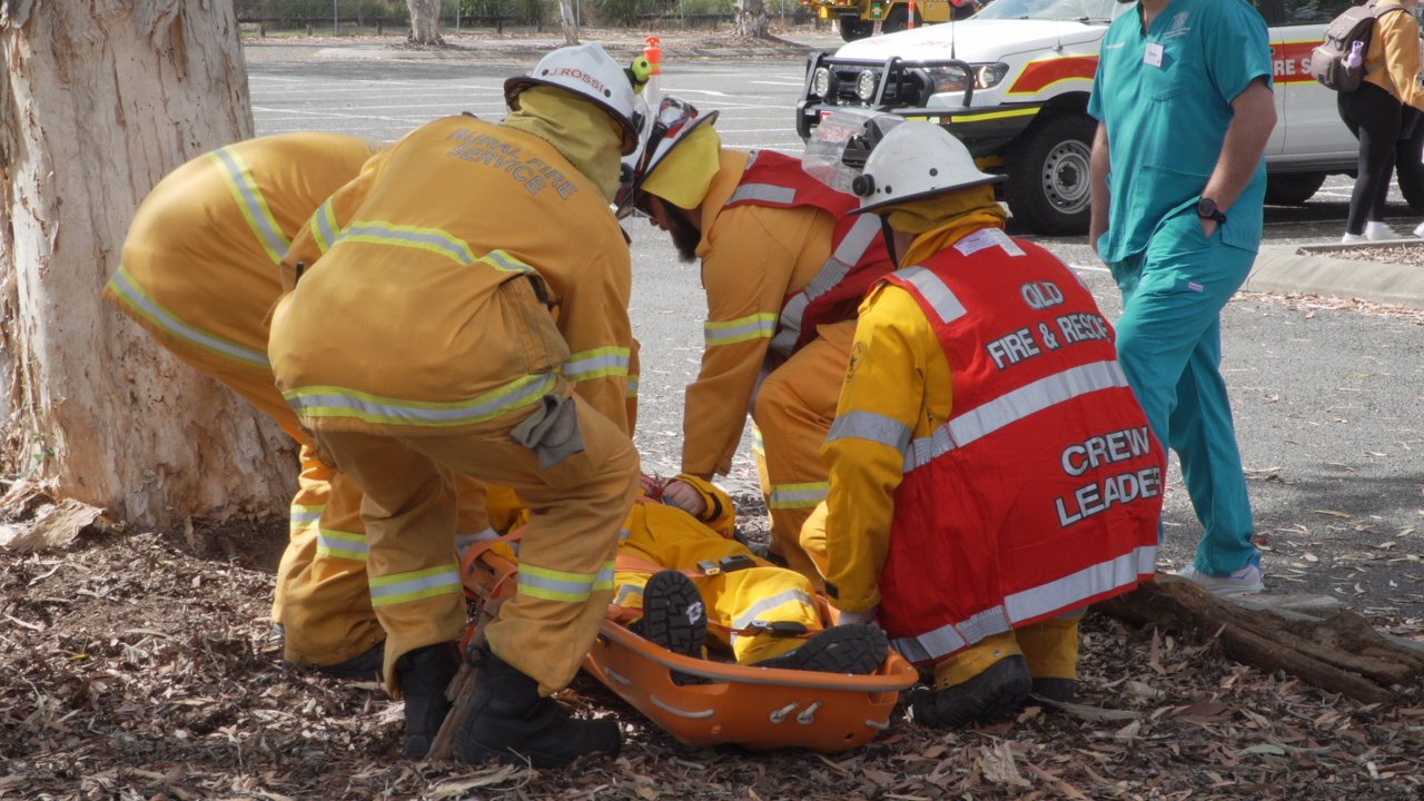 Firefighters provide emergency medical assistance to a person on a stretcher outdoors, near a tree, with an ambulance and a person in scrubs nearby.