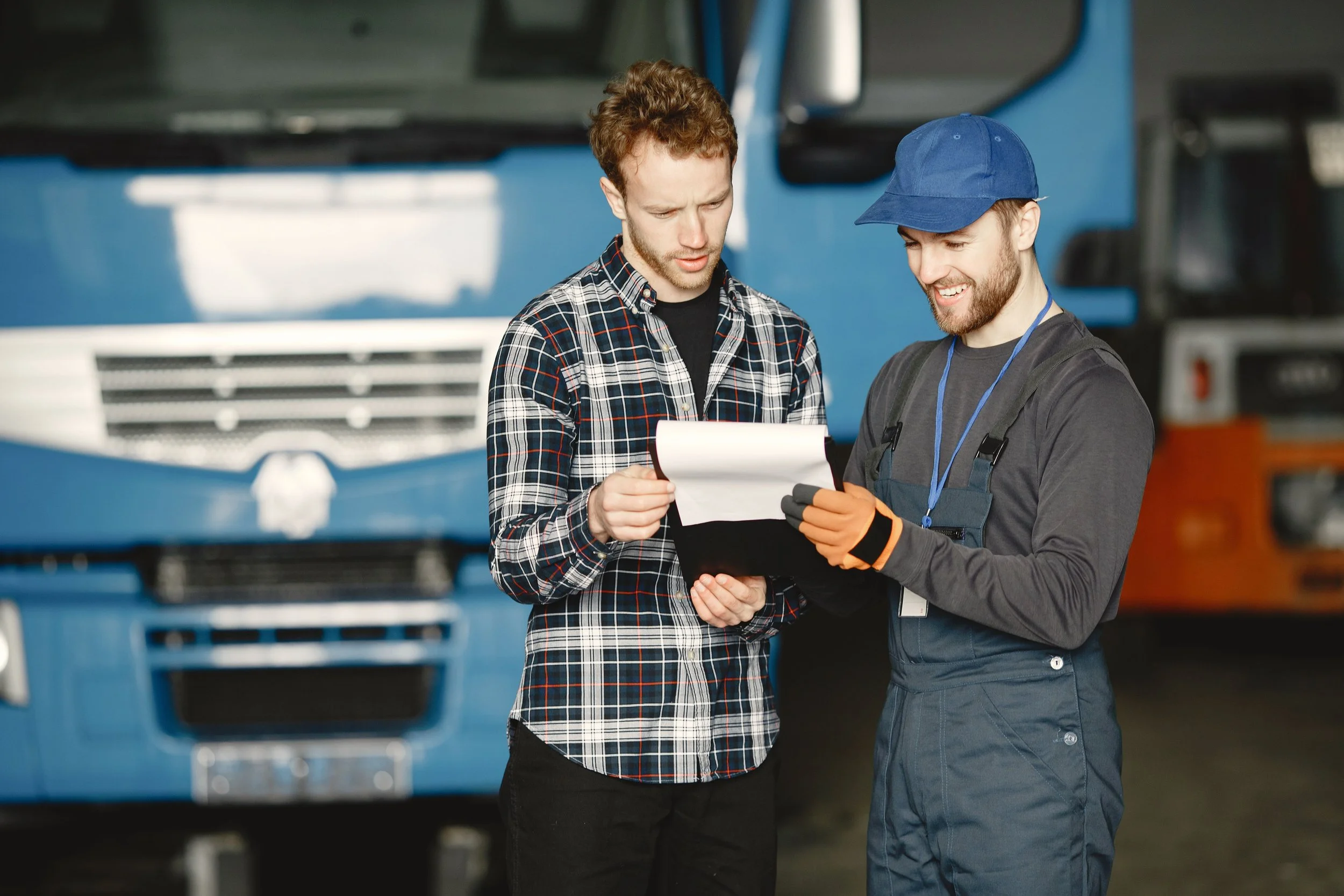Two men are standing in front of a large blue truck, looking at a clipboard and smiling. One man is wearing a checkered shirt and the other is in work overalls and a blue cap.