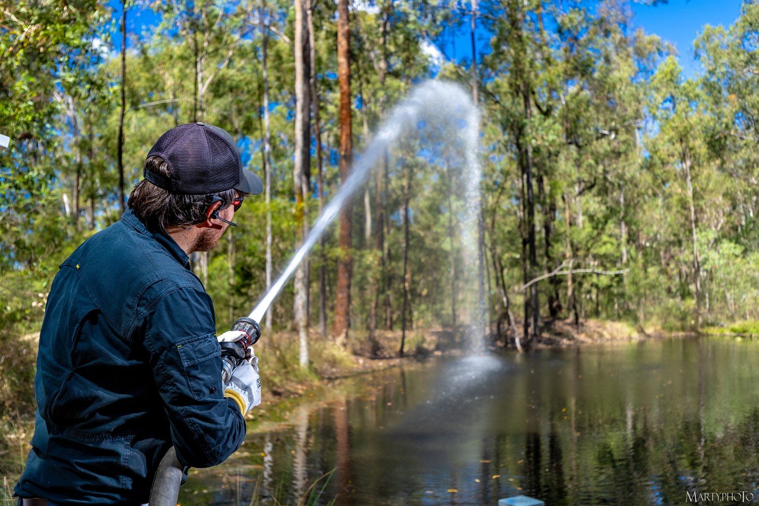 A man in a dark blue shirt and black cap, wearing a headset, is spraying water from a hose into a body of water in a wooded area with green trees and a blue sky.