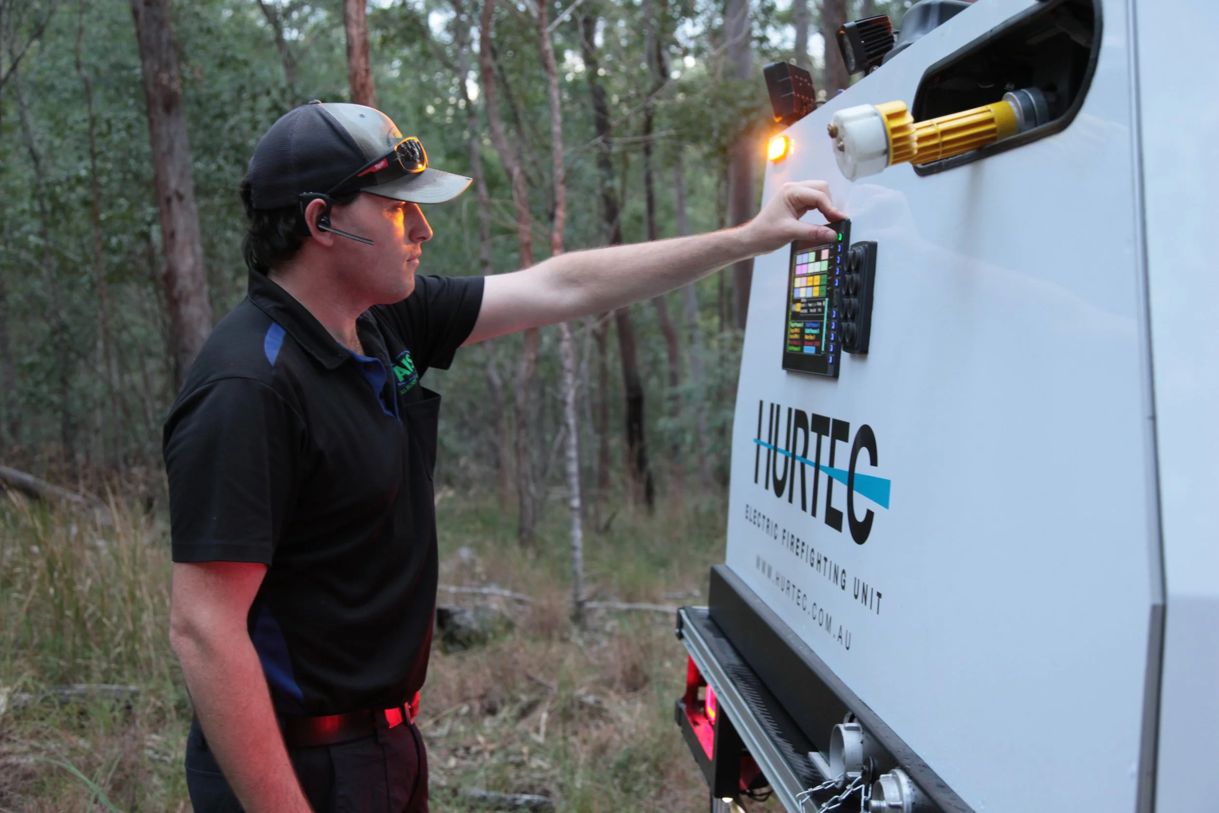 A man wearing a headset, sunglasses, and a black shirt stands outdoors in a forested area, operating Hurtec electrical firefighting equipment in a slip-on firefighting back.