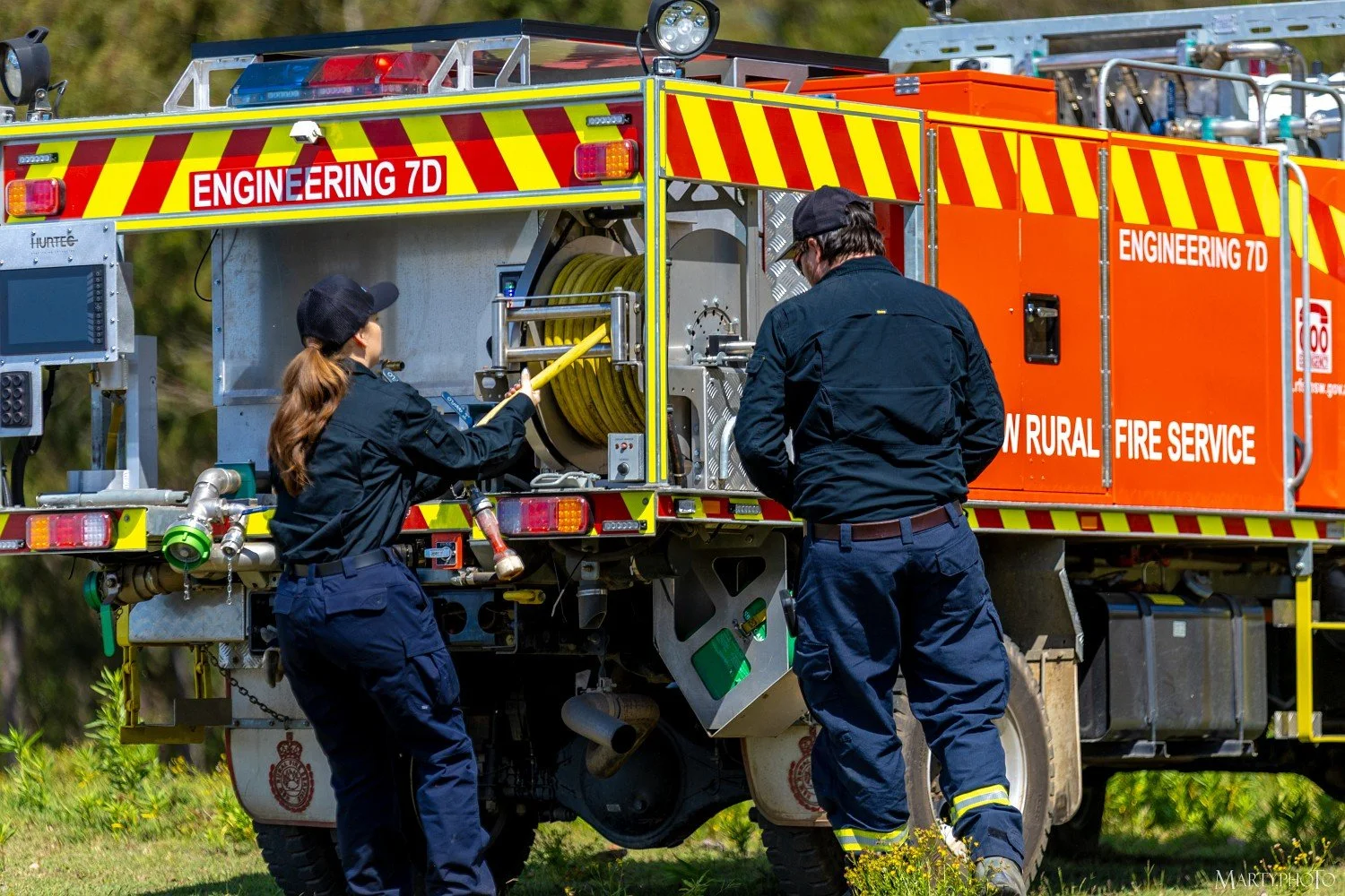 Two firefighters working on a fire truck fitted with a Hurtec electric firefighting pump and Hurtec FleetCon software, with one pulling a yellow hose and the other inspecting equipment.