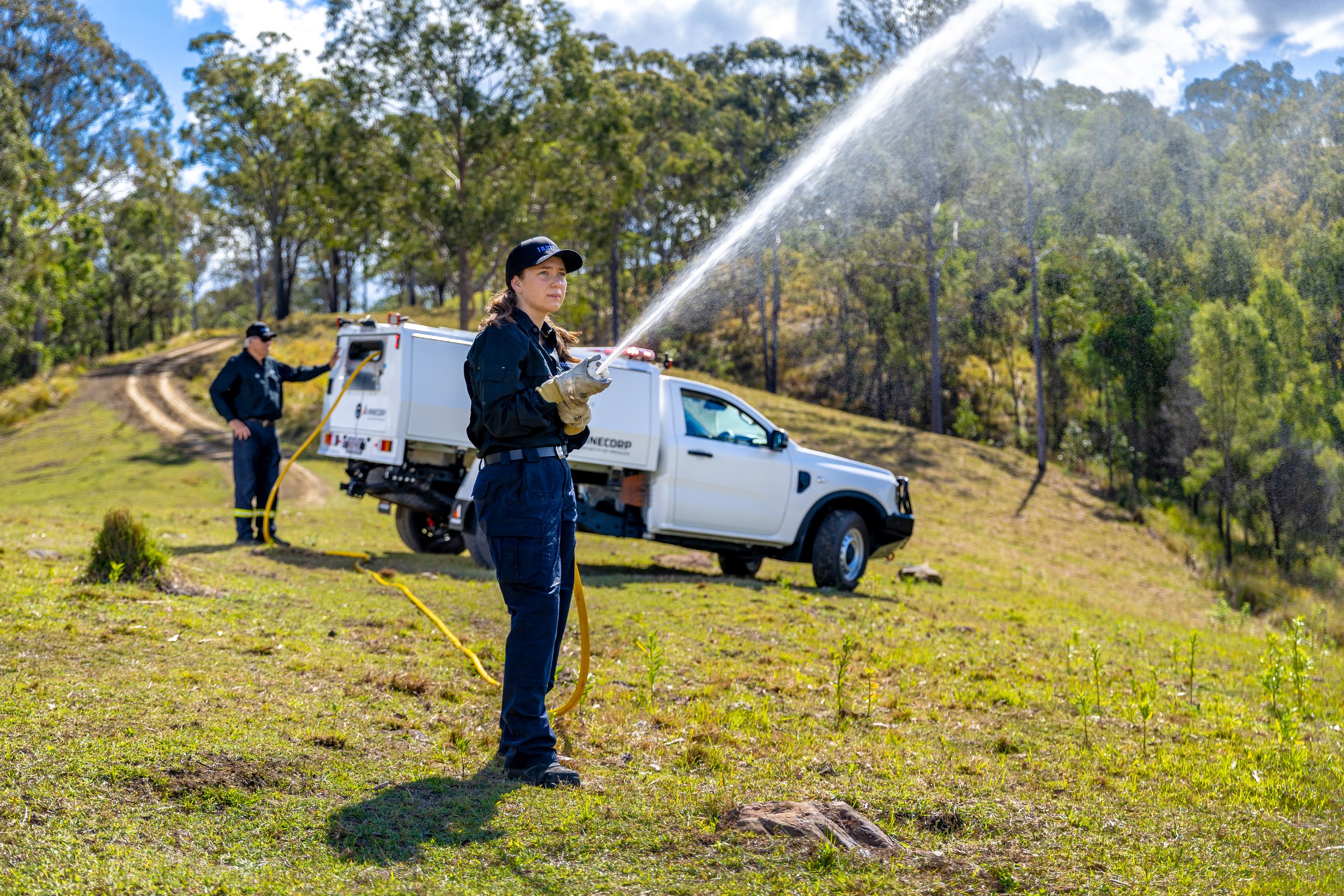 Two firefighters in uniform spray water into the air while standing on grassy terrain near a white utility vehicle with trees and a partly cloudy sky in the background.