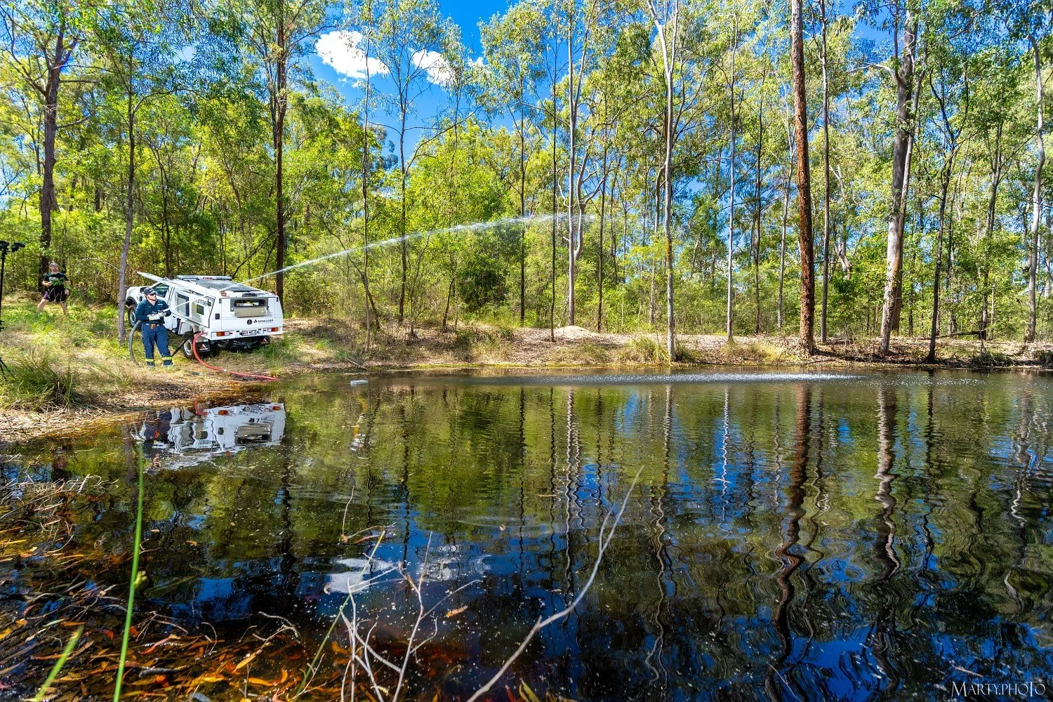Firefighters spraying water on a pond in a wooded area during daytime.