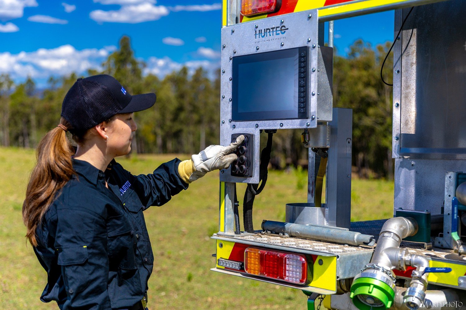 A woman in a black uniform and cap operating a Hurtec FleetCon firefighting pump control panel on a piece of industrial equipment outdoors, with trees and a blue sky in the background.