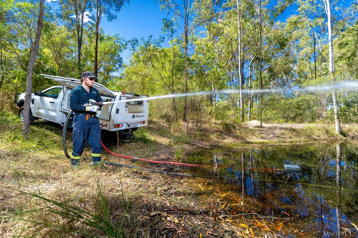 A firefighter in navy uniform and yellow gloves spraying water from a hose into a pond in a wooded area, with a white emergency vehicle parked nearby.
