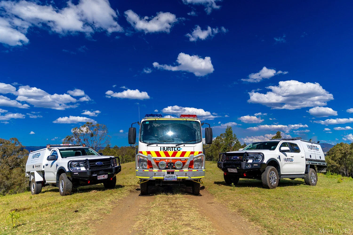 Three emergency rescue vehicles parked on grass outdoors under a blue sky with clouds. The center vehicle is a large fire truck, flanked by two pickup trucks with rescue equipment, all marked with 'HURTEC' and emergency lights on top.