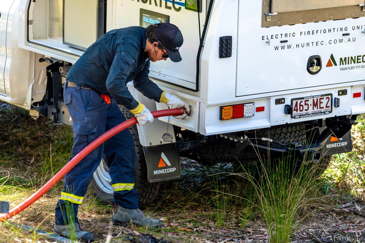A worker in navy uniform and yellow gloves is connecting a red hose to a white emergency vehicle labeled 'Electric Firefighting Unit.' The vehicle is parked on grassy terrain with some foliage.