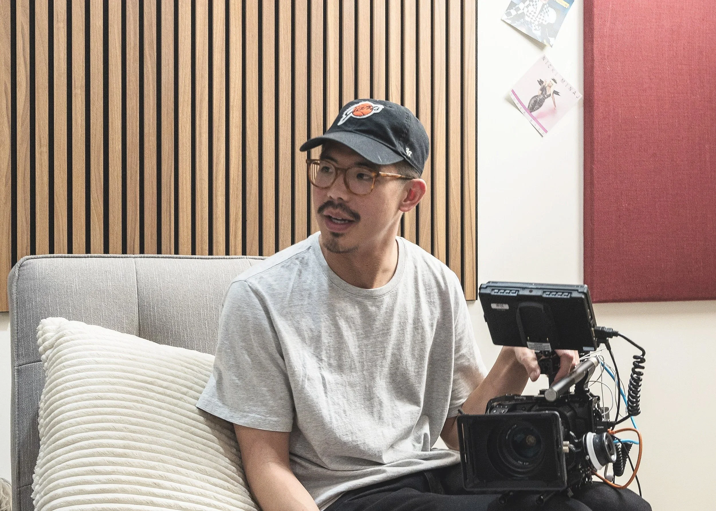 A professional filmmaker, videographer, and documentarian, holding a camera, sitting on a gray couch in a room with wooden paneling and polka dot posters on the wall.