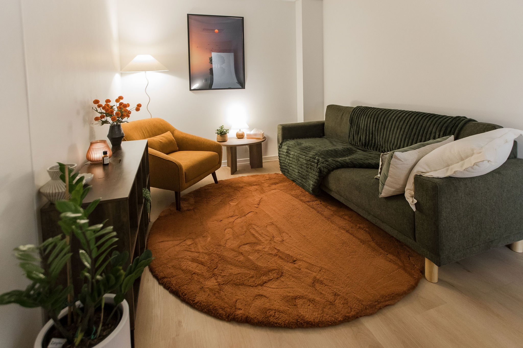 A cozy living room with a green velvet sofa, an orange armchair, a round wooden side table, and a dark wooden console table. The room features a large, oval, brown shag rug on light wooden flooring, a white wall with a landscape photograph, and various decorative plants and lamps.