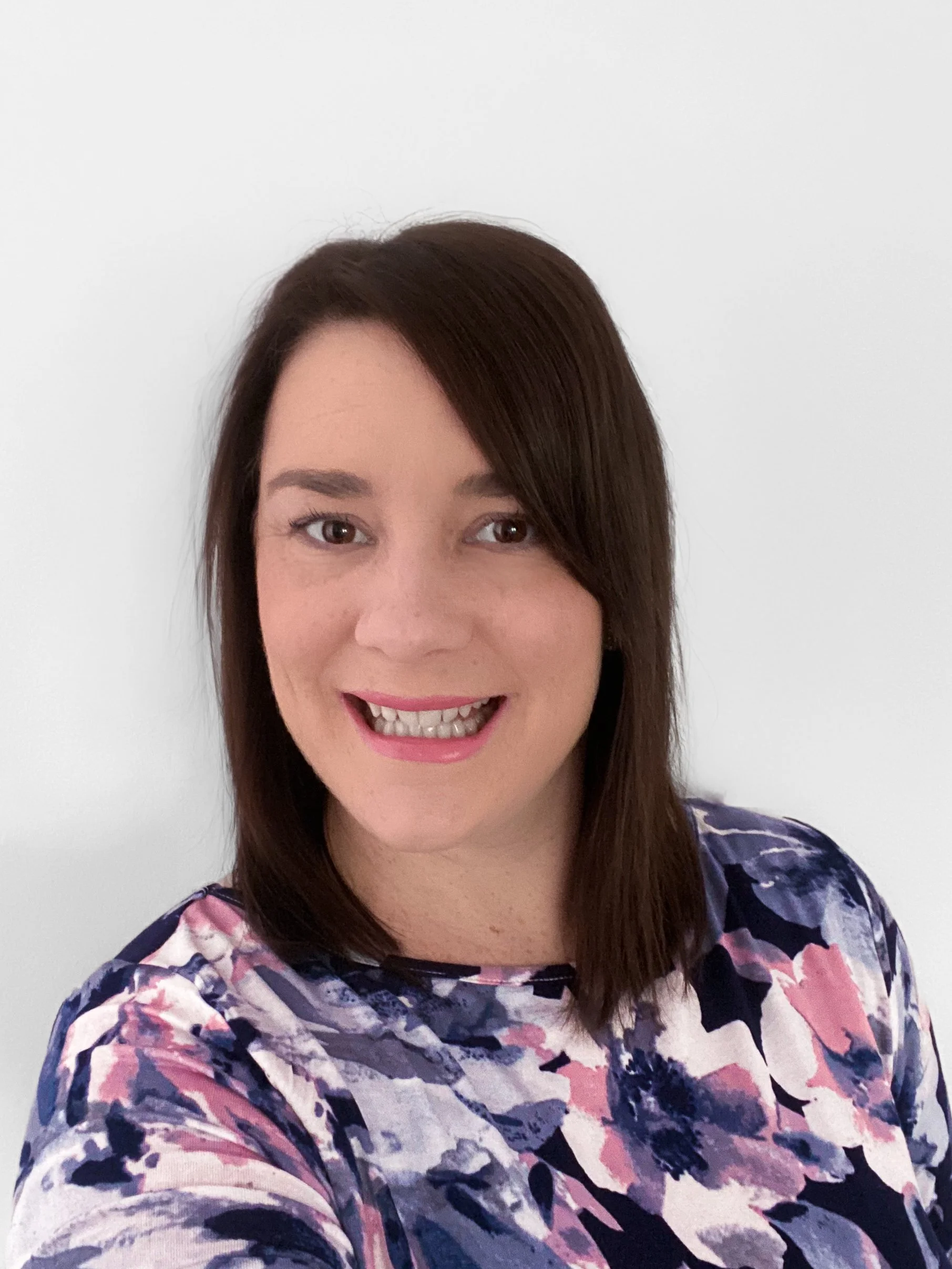 Close-up selfie of a woman with shoulder-length brown hair and a floral-patterned top, smiling against a plain white background.