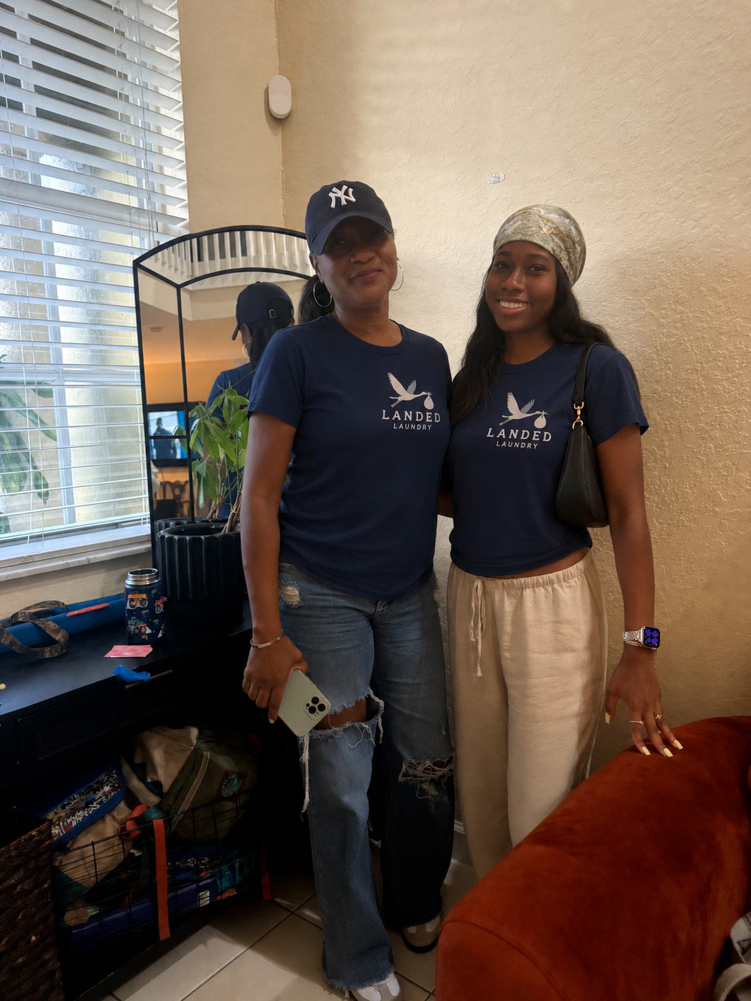 Two women standing indoors near a window, both wearing navy blue T-shirts with a logo for Landed Laundry. The woman on the left is holding a phone and wearing a baseball cap, while the woman on the right is wearing a headscarf and a smartwatch. They are smiling for the photo.