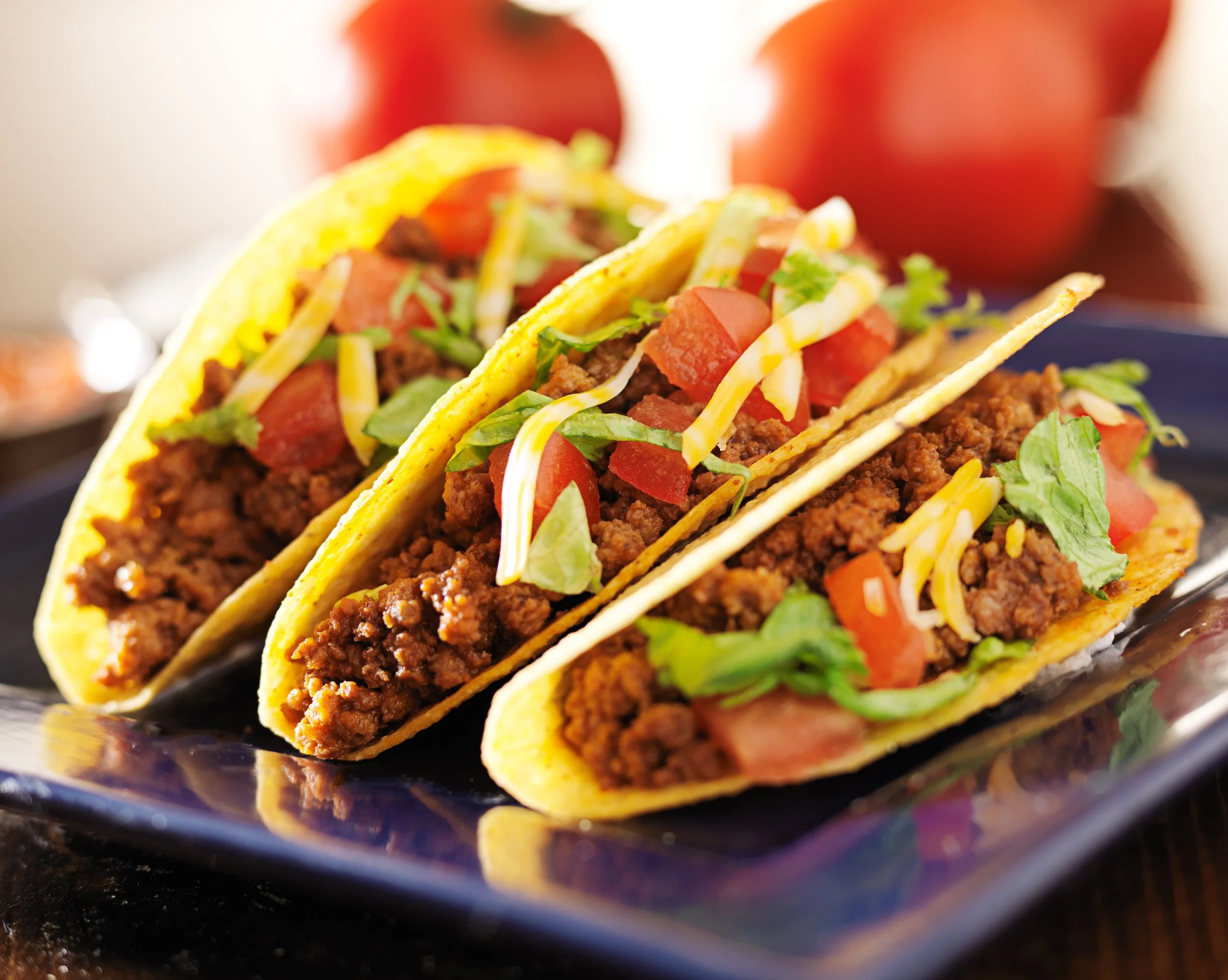 Close-up of three tacos filled with ground beef, chopped tomatoes, shredded cheese, and lettuce on a black plate.