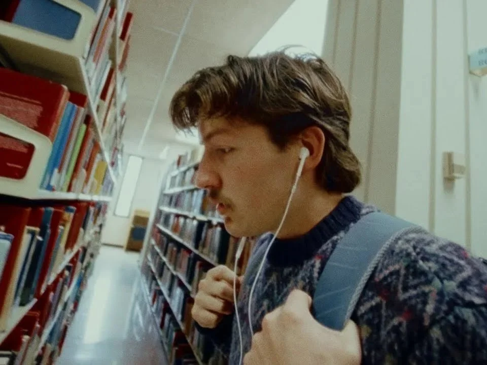 Young man in a library with glasses, wearing a sweater, carrying a backpack, and listening to headphones.