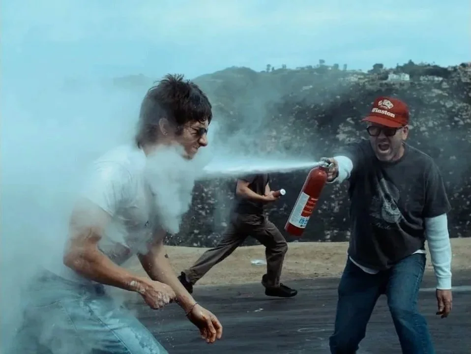 Two men fighting with a fire extinguisher on a windy outdoor landscape, one being sprayed and the other dodging the spray.