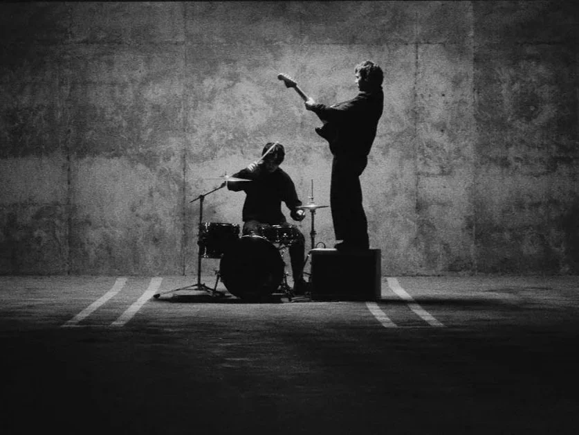 Two musicians performing; one on drums and one on guitar, in a minimalistic setting with a concrete wall background, black and white image.