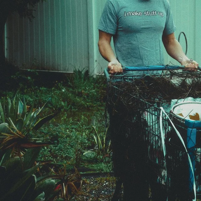 Person in a blue t-shirt holding a wire basket filled with tangled vines or weeds in a garden