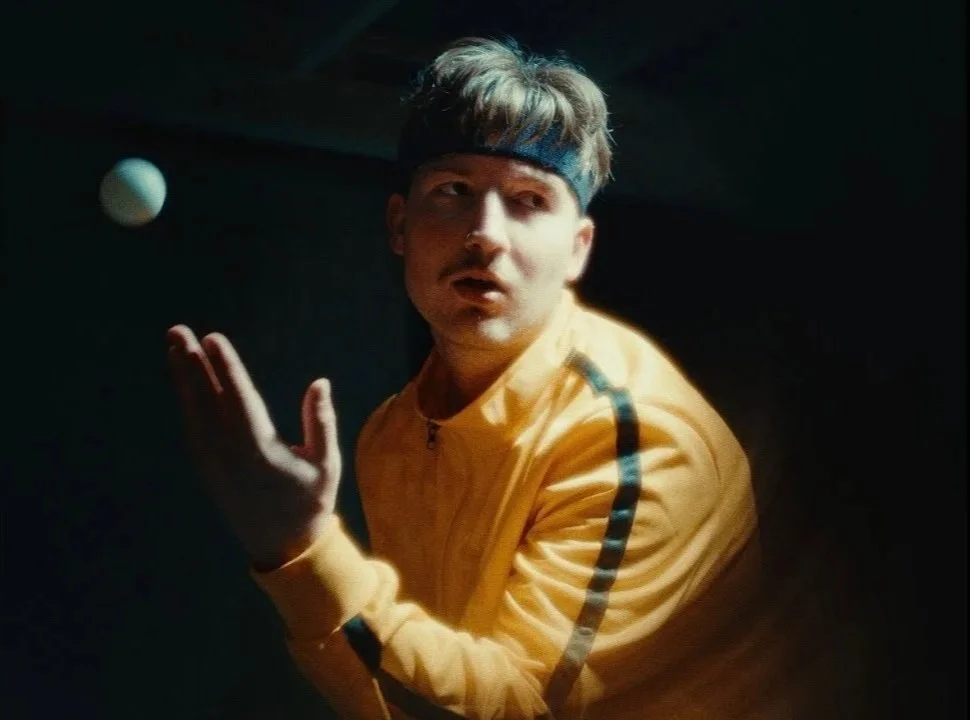 A young man wearing a yellow jacket and a dark headband is preparing to hit a tennis ball, which is visible in the air near him, in a dimly lit environment.