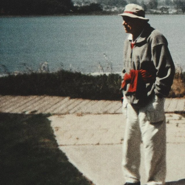 A person wearing a wide-brimmed hat, gray jacket, and white pants walking outdoors near a body of water with a cloudy sky in the background.