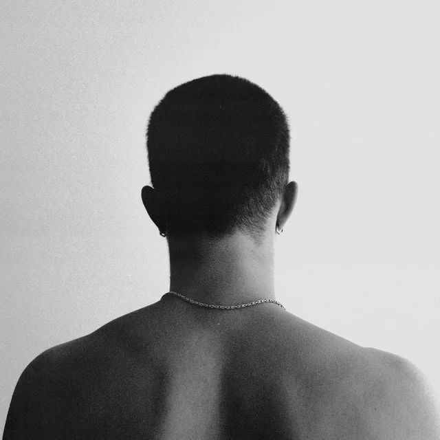 Back view of a person with short hair, wearing earrings and a chain necklace, against a plain background.