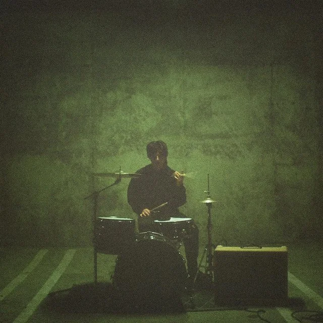A person playing drums in a dimly lit, greenish environment, with an electric guitar and amplifier nearby.