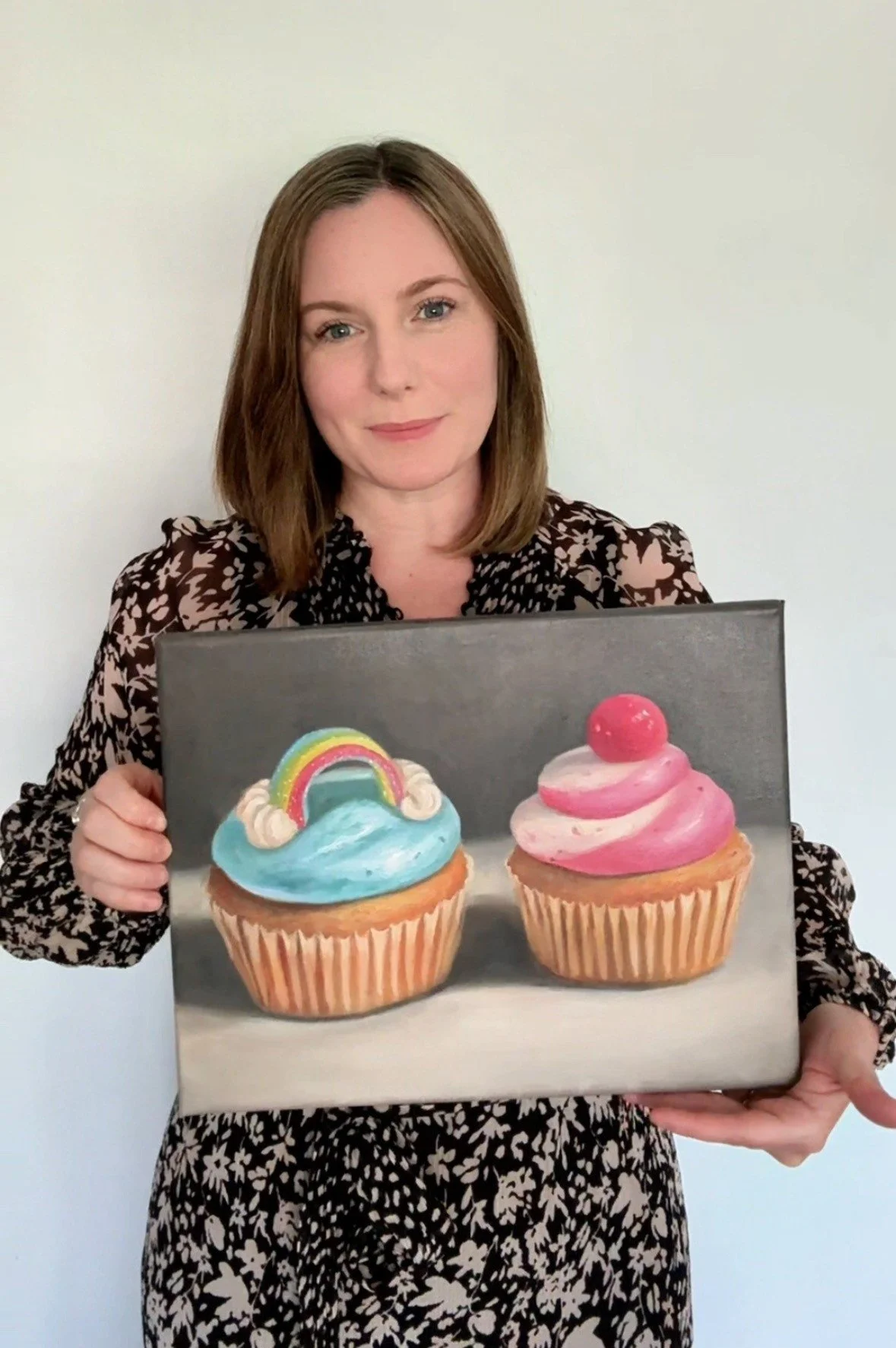 Dani Giles holding a painting of two cupcakes, one with rainbow and clouds and the other with pink frosting and a gumball on top.