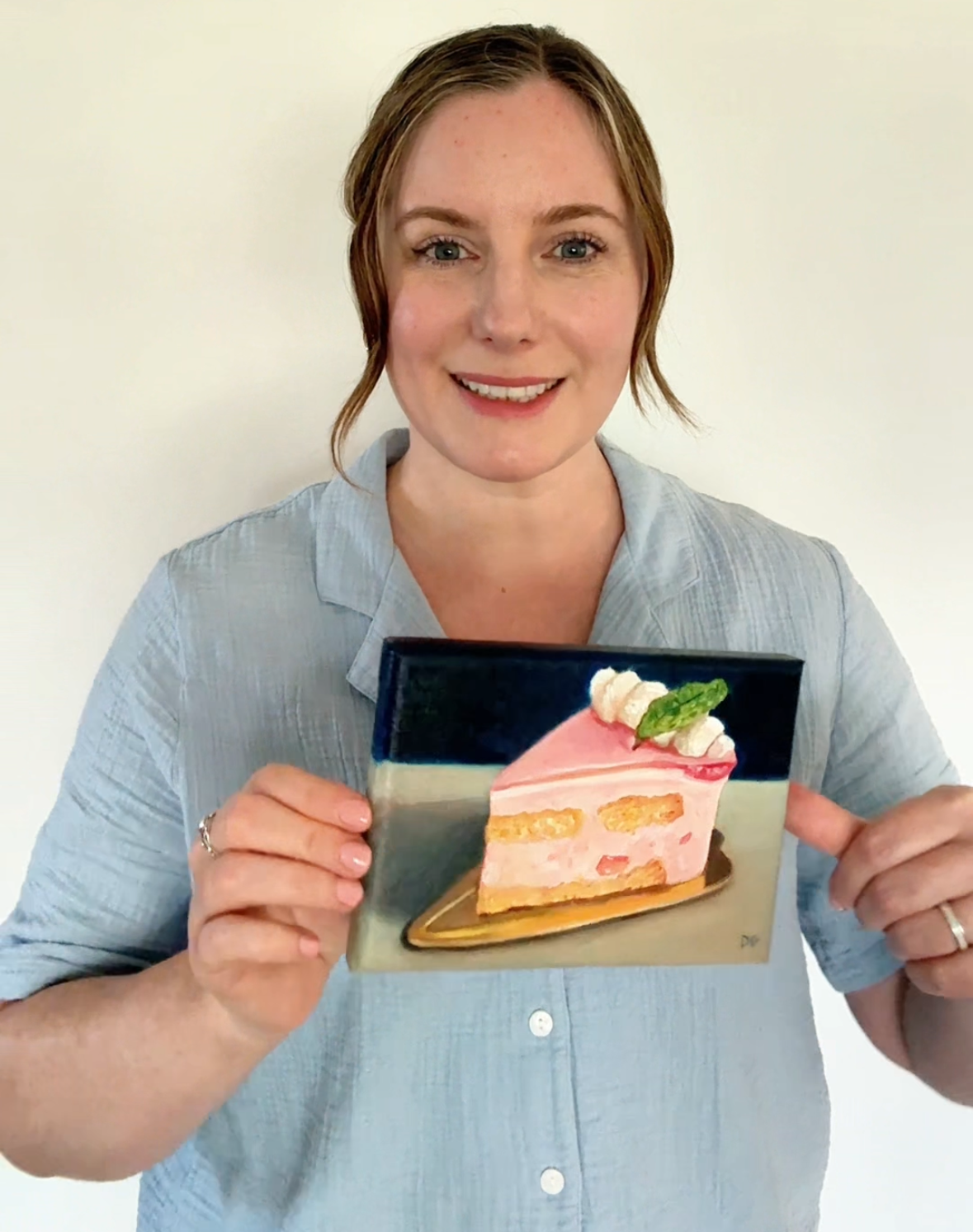 Dani holding an oil painting of a slice of pink cake.