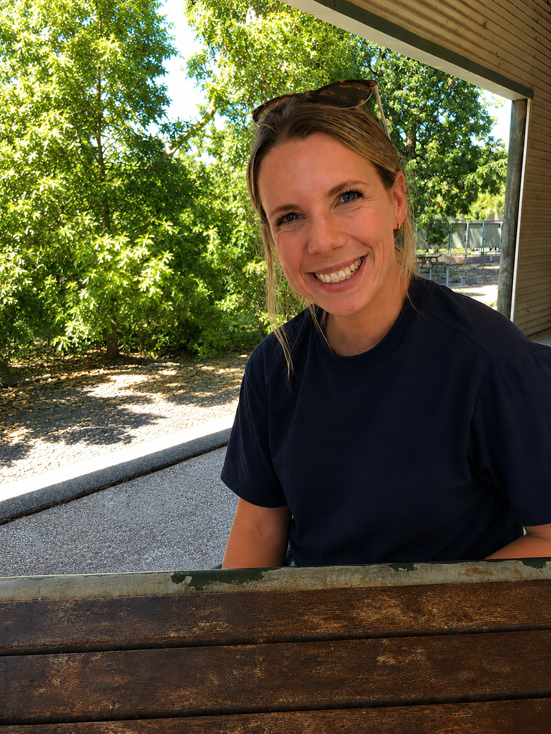 A smiling woman with blonde hair, sitting outdoors at a wooden table, with green leafy trees in the background, on a sunny day.