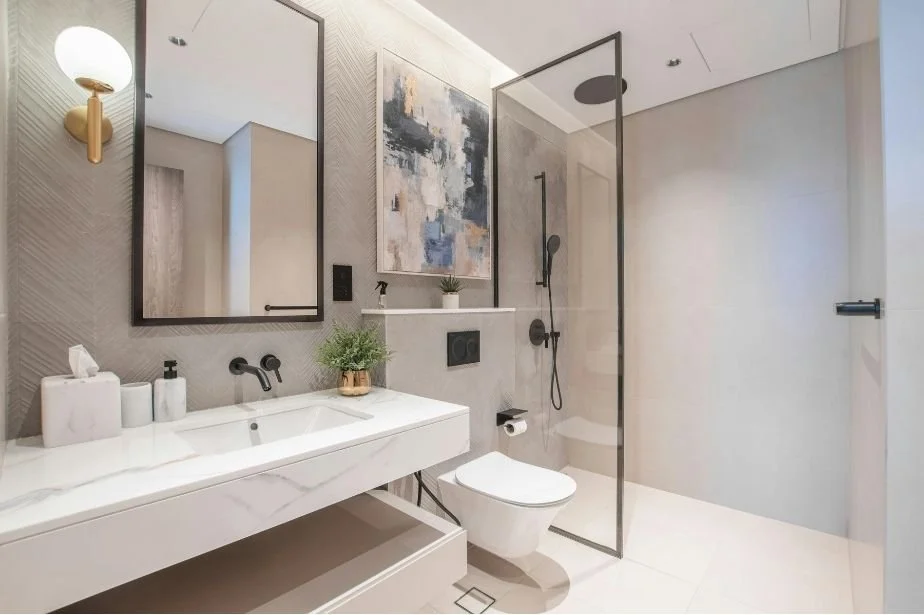 Modern bathroom with a floating white sink, large wall mirror, art piece, plant, wall-mounted black faucet, toilet, and level access shower with half glass screen.