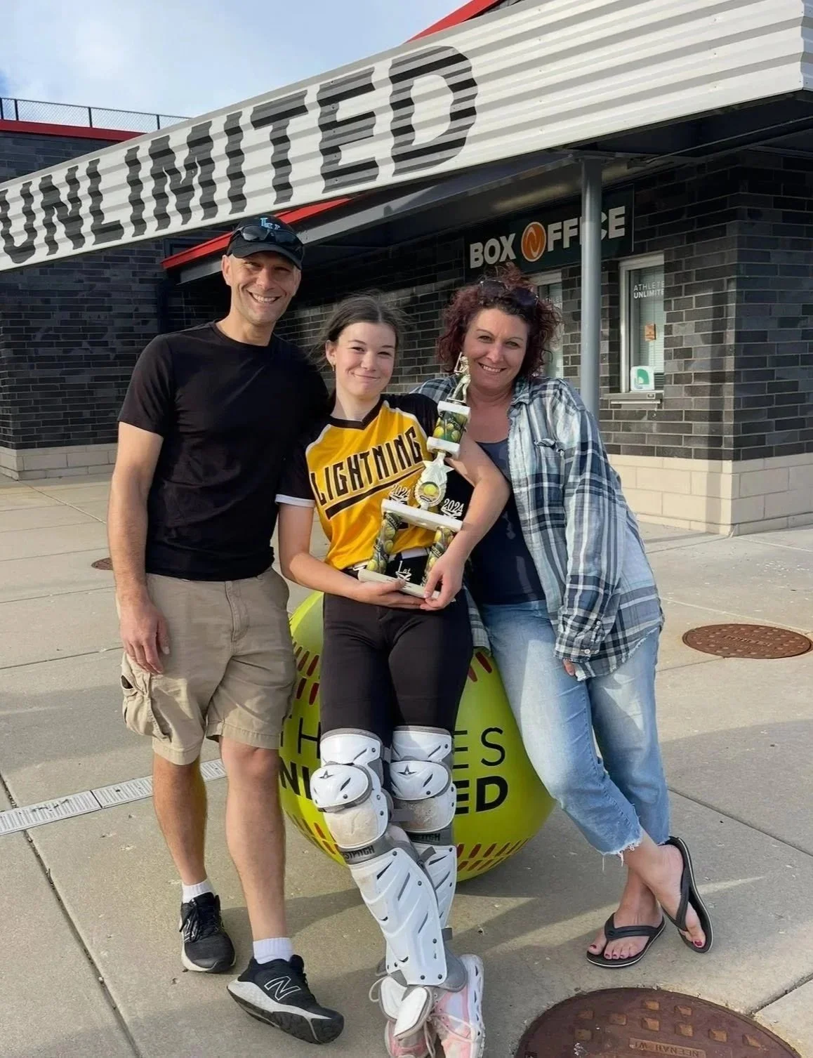 Three people standing outside a building with a sign that says 'UNLIMITED' and a window that says 'BOX OFFICE.' The person in the middle is a young girl wearing sports gear and holding a large trophy, with roller hockey goalie pads. The two adults on either side are smiling; the person on the left is a man in a black t-shirt and beige shorts, and the person on the right is a woman in a plaid shirt and jeans.