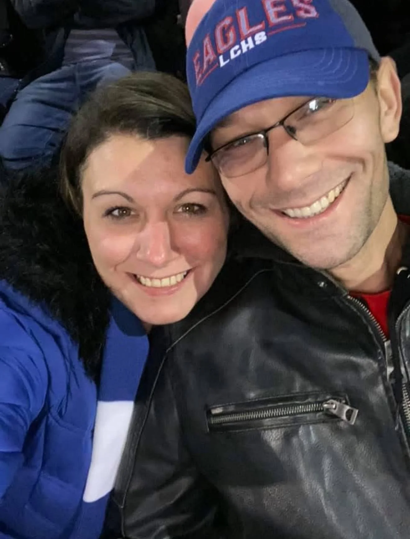 A smiling man and woman taking a selfie together. The woman has long dark hair and is wearing a blue jacket. The man is wearing glasses, a blue cap with red and white lettering, and a black leather jacket.