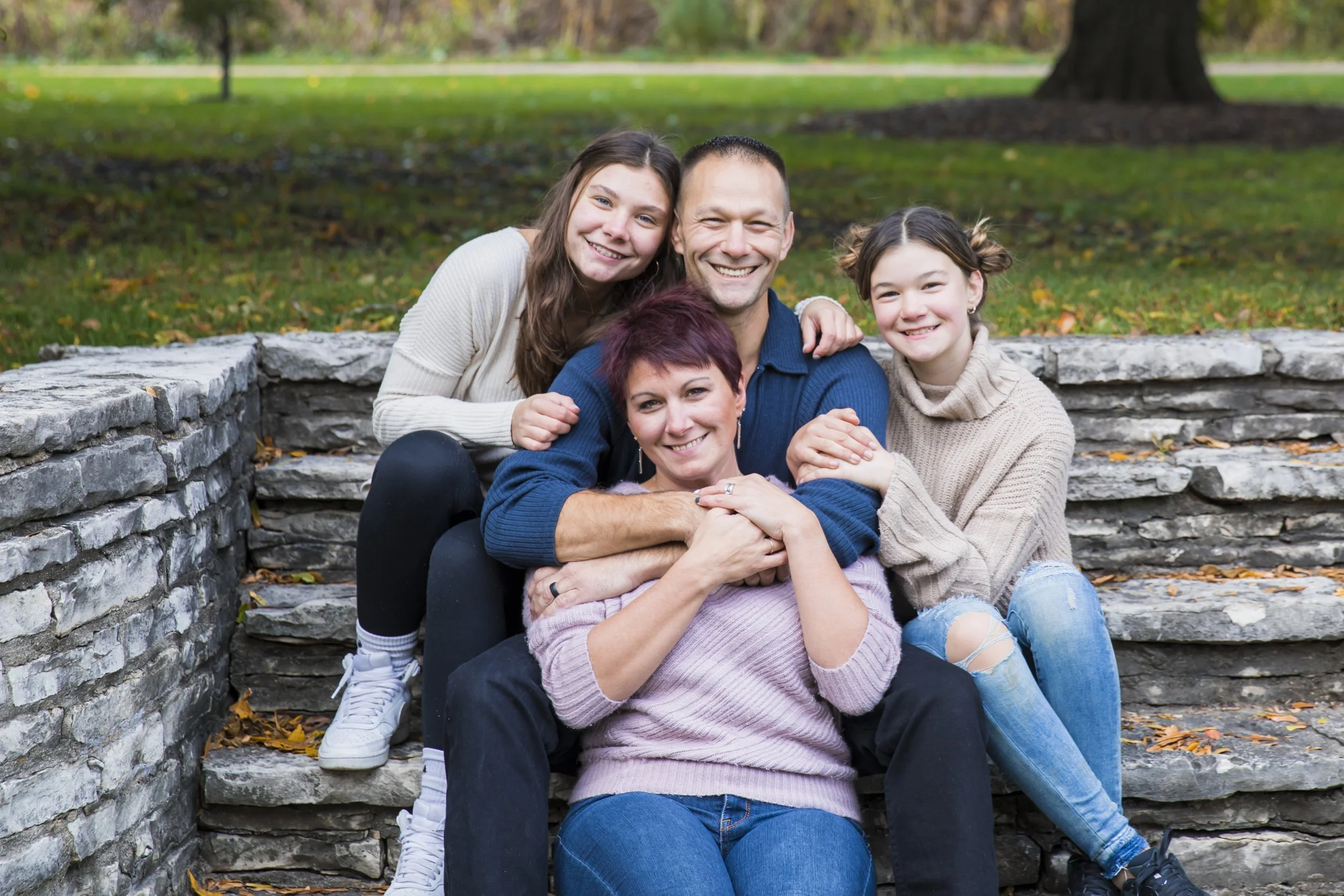 A family of five sitting on stone steps outdoors in a park, smiling and hugging each other on a fall day, with a tree and grassy area in the background.