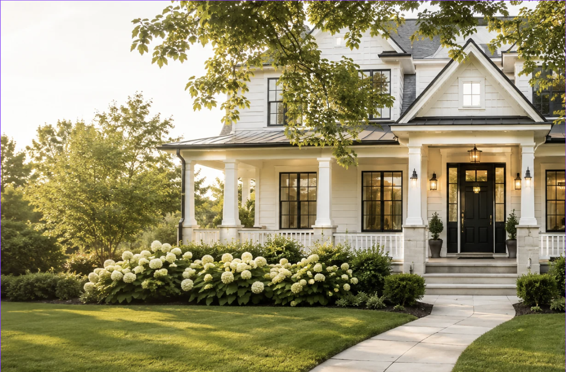 A white, two-story house with a front porch, black door, and large windows. The house is surrounded by a well-maintained lawn, lush green trees, and white flowering bushes, with steps leading up to the front door and outdoor lanterns on either side.
