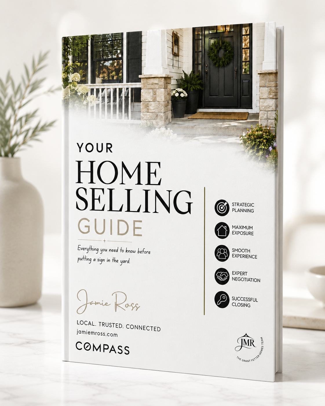 A home selling guidebook with a picture of a front porch with a black door and wreath, potted plants, and flowers, standing on a white table next to a beige vase with green foliage in the background.