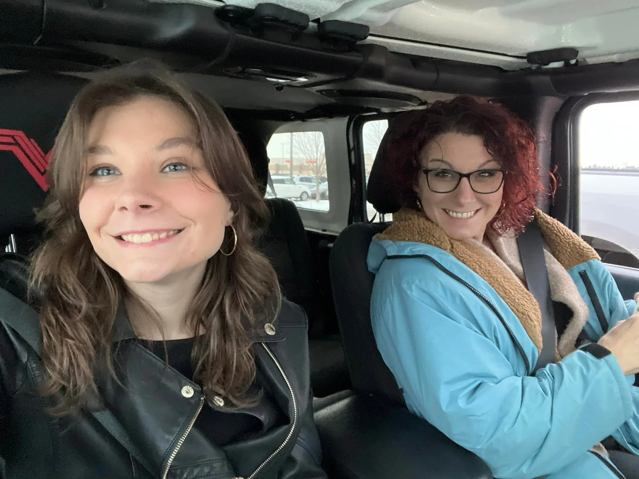 Two women sitting in the front seats of a vehicle, smiling at the camera. The woman on the left has long, wavy brown hair and wears a black jacket, while the woman on the right has curly red hair, glasses, and wears a blue jacket with a tan collar. The background shows a parking lot through the vehicle's windows.