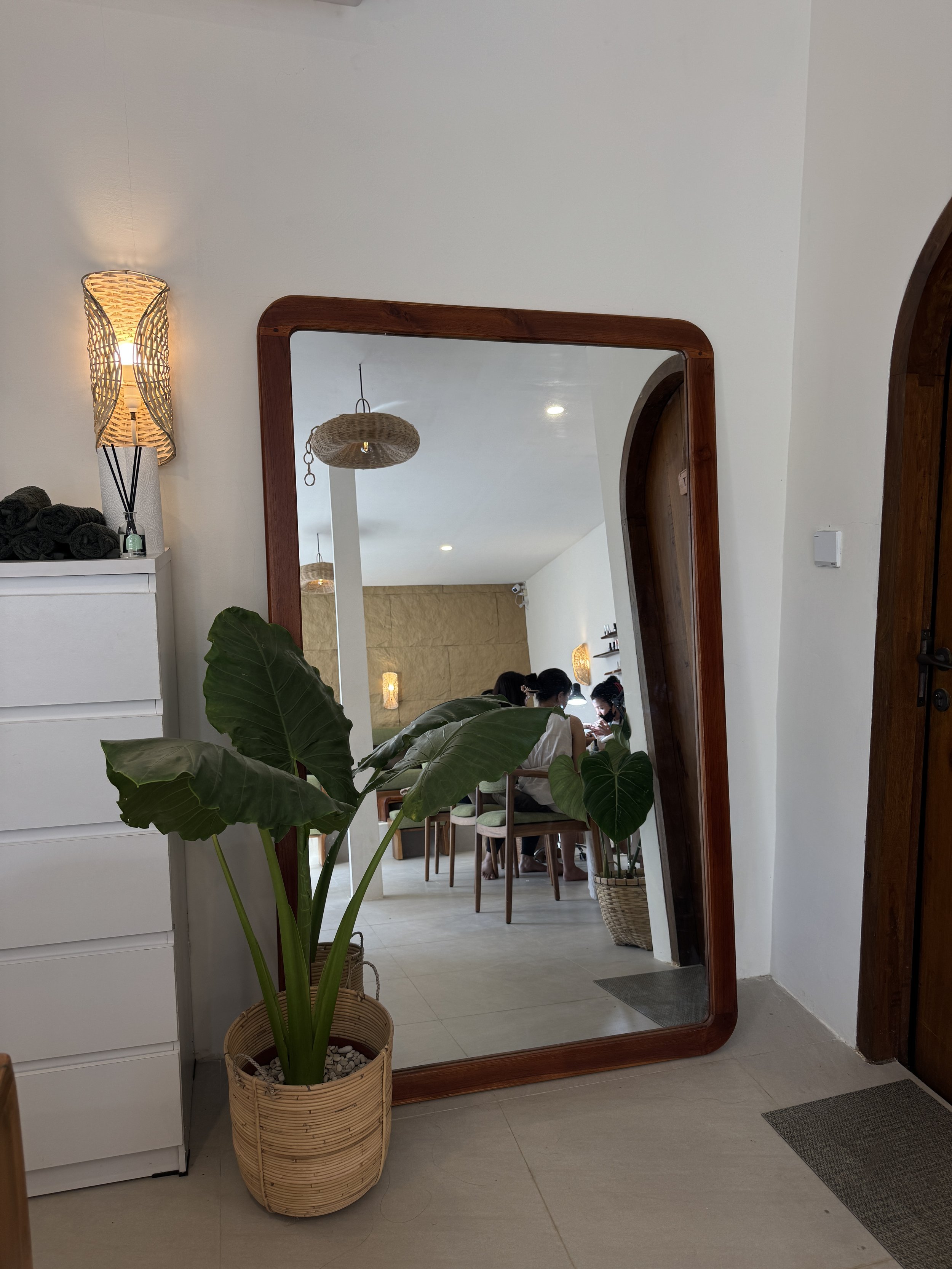 An interior space with a large wooden framed mirror leaning against the wall, a potted plant with large green leaves in front of it, a white chest of drawers with rolled black towels on top, and a wall-mounted lamp with a woven shade. In the reflection, there are several people sitting at a table inside a cozy room with beige walls and hanging woven light fixtures.