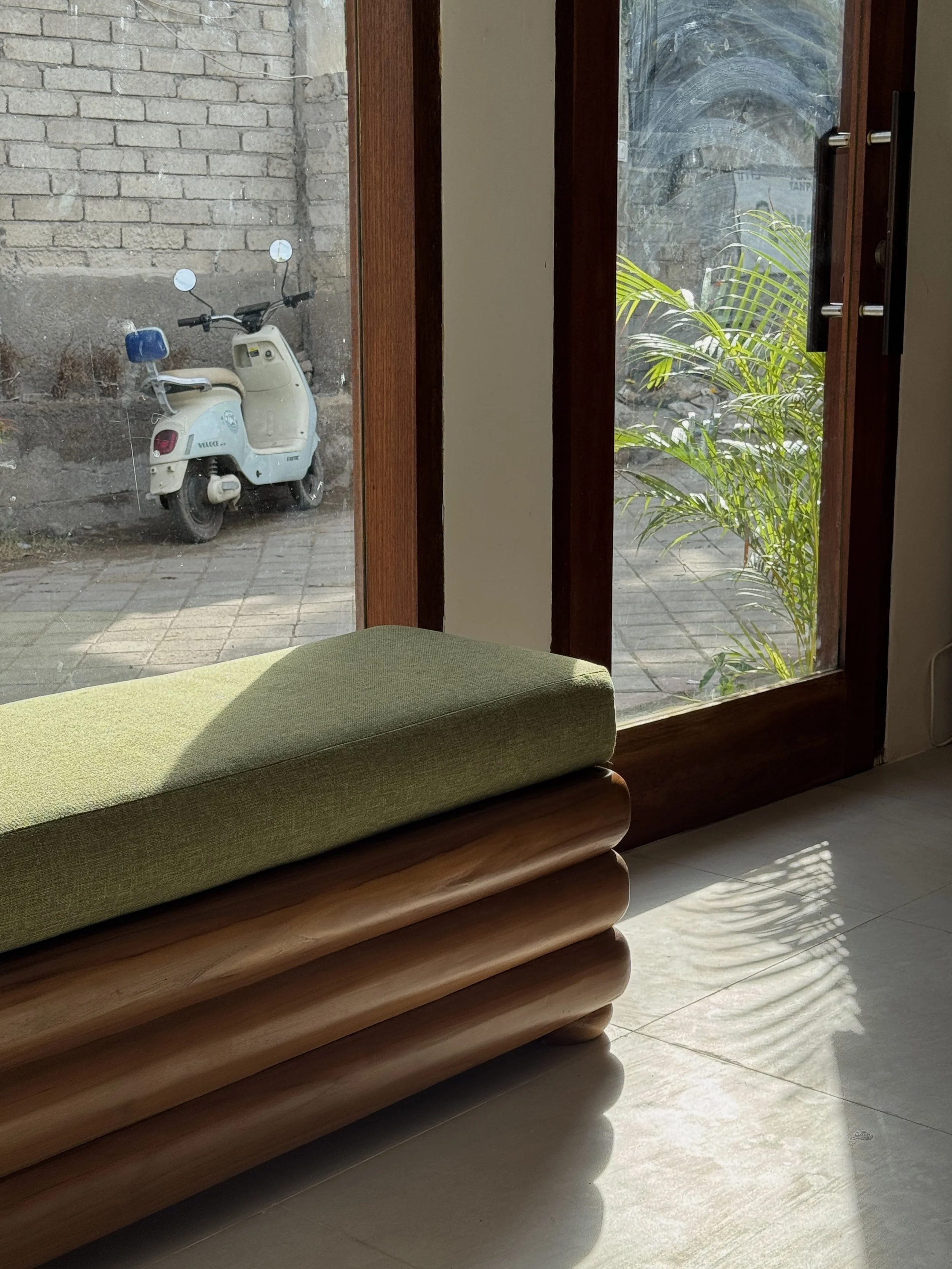 View of a wooden bench with a green cushion inside a room, looking out through a glass door to a patio area with a white scooter parked against a brick wall and some green plants outside.