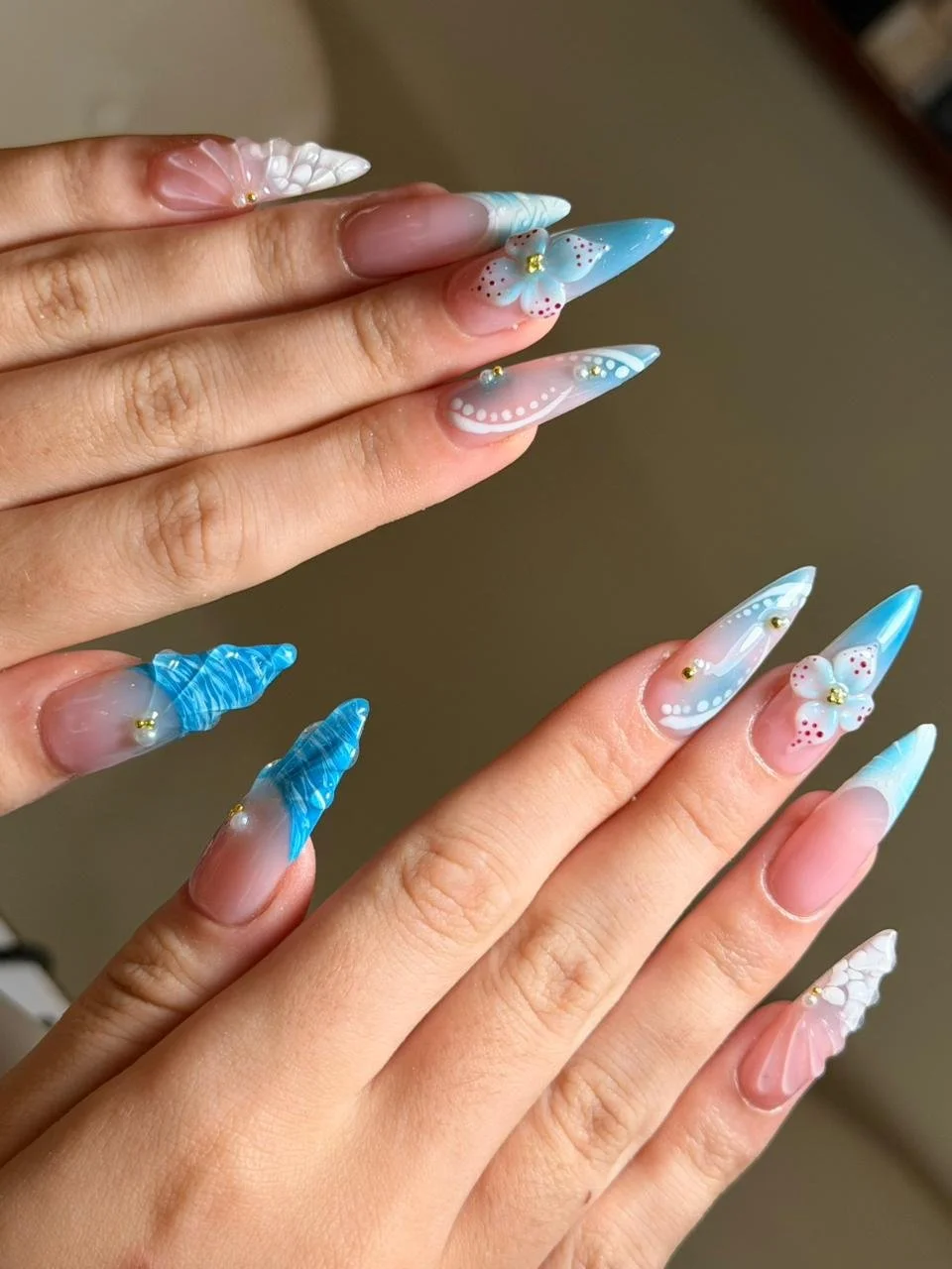 Close-up of hands with long, elaborately decorated acrylic nails in pastel colors, featuring floral designs, rhinestones, and textured patterns.