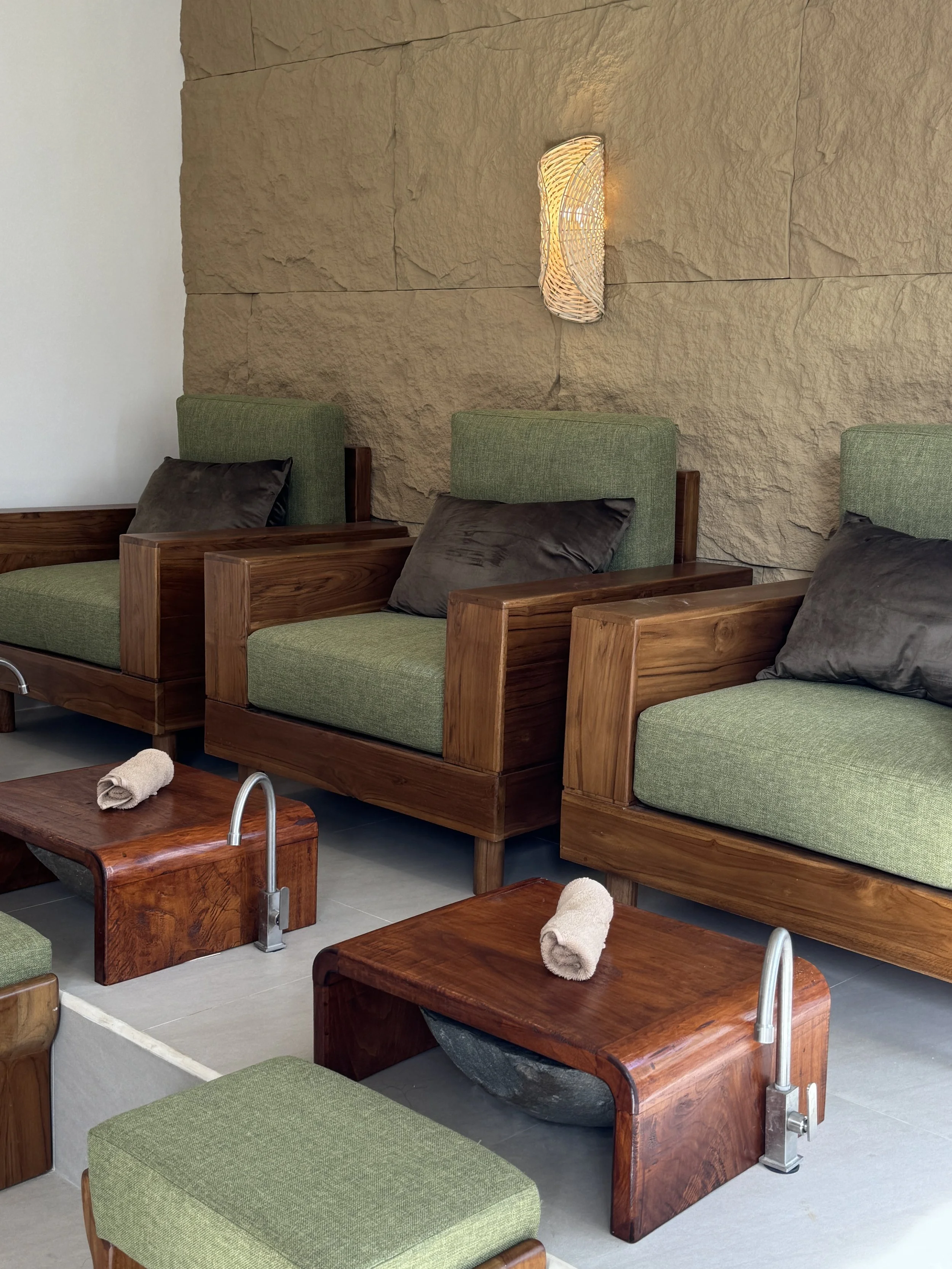 A spa or massage room with wooden beds and green upholstered cushions, some with dark pillows. Small towel rolls are placed on the beds. There are large stone tiles on the wall with a modern light fixture beside them.